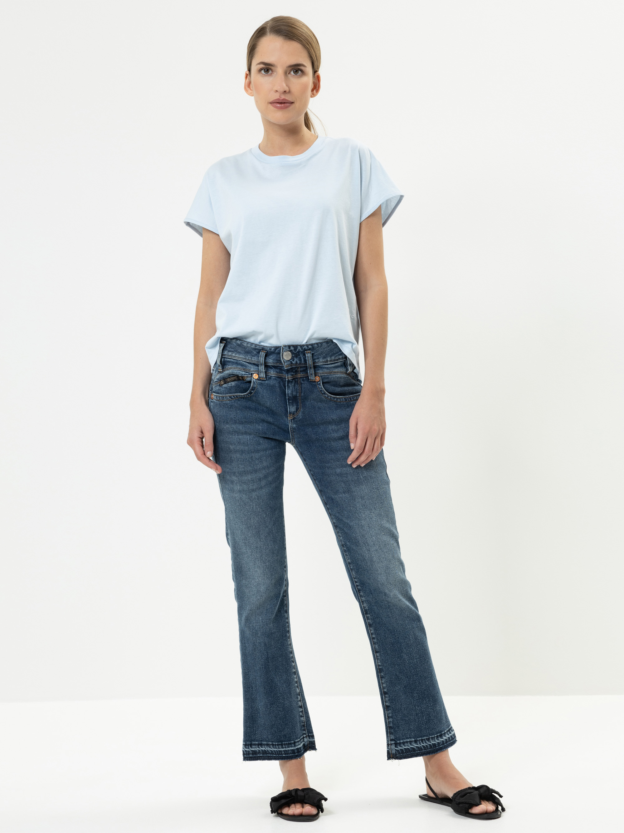 Eine Frau steht vor einem schlichten weißen Hintergrund und trägt ein hellblaues T-Shirt, blaue Jeans mit geradem Bein und schwarze Slide-Sandalen mit Schleifen. Sie hat ihr Haar zurückgebunden und schaut mit neutralem Blick direkt in die Kamera.