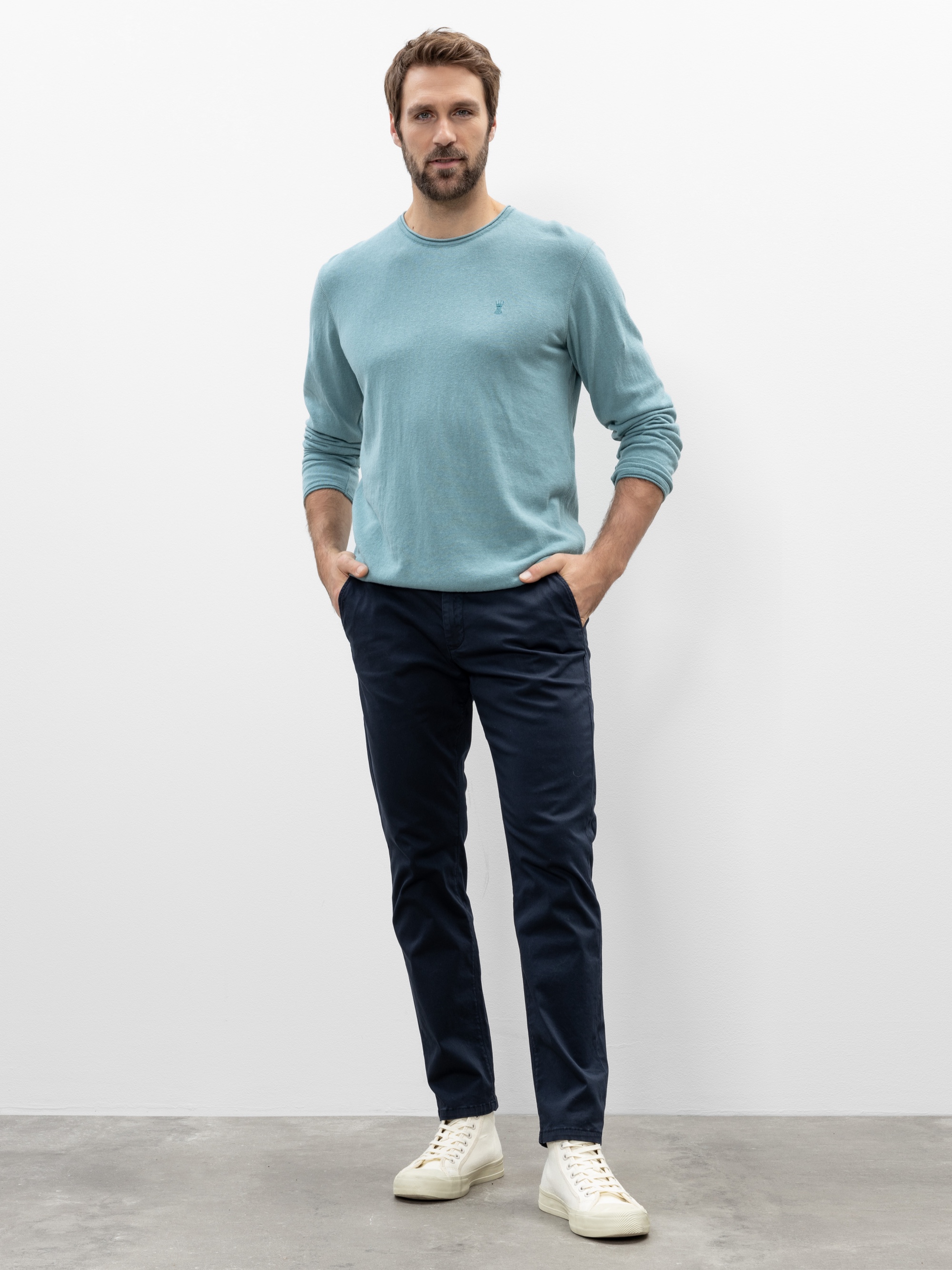 Ein Mann steht mit einem hellblauen langärmeligen Hemd, einer dunkelblauen Hose und weißen Turnschuhen an einer schlichten weißen Wand und hat die Hände in den Taschen.