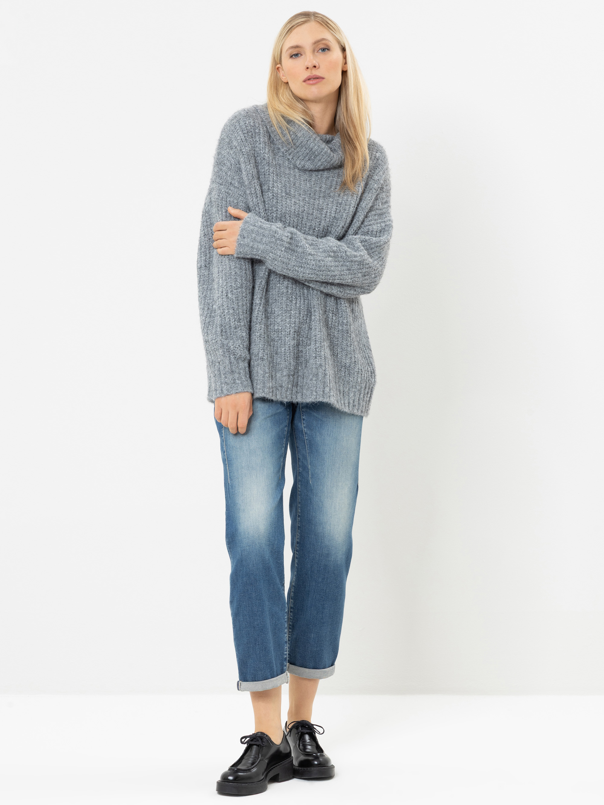 Vor einem weißen Hintergrund steht eine Frau, die einen lockeren grauen Rollkragenpullover, eine blaue Jeans mit Umschlag und schwarze Schuhe trägt. Sie hat glattes blondes Haar und nimmt eine entspannte Haltung ein, wobei sie mit einem Arm den gegenüb