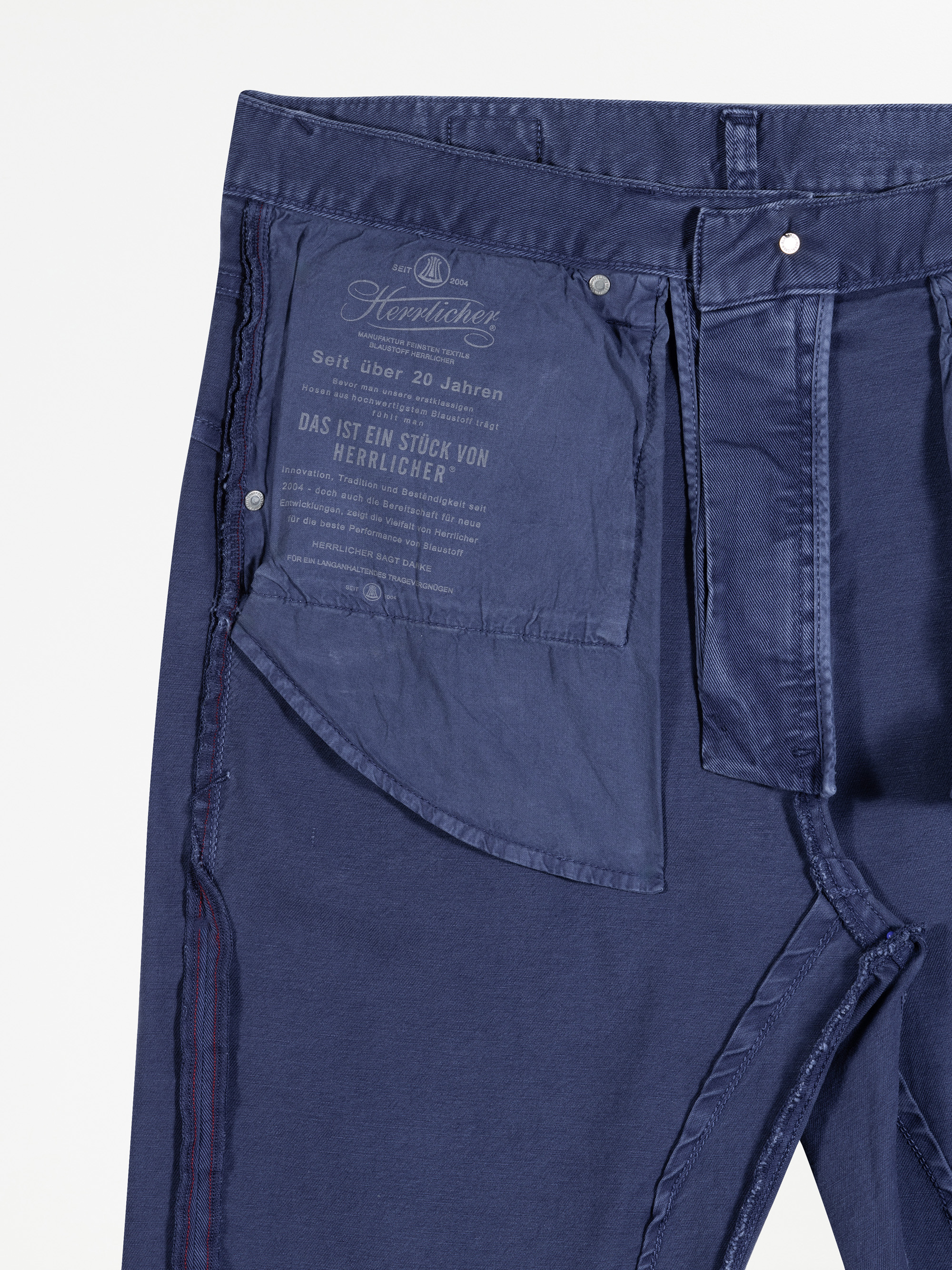 Die dunkelblaue Hose ist von innen nach außen gekrempelt und zeigt das Innenfutter der Tasche mit aufgedrucktem Text und Branding, einschließlich des Satzes "Das ist ein Stück von Herrlicher". Die Hose hat einen Knopfverschluss und sichtbare Nähte.