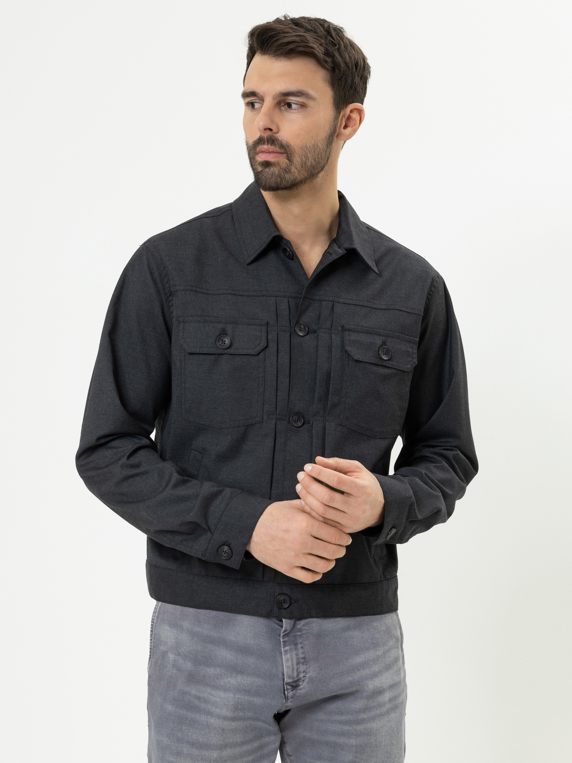 Ein Mann mit kurzen dunklen Haaren und einem Bart trägt eine dunkelgraue, geknöpfte Jacke mit Brusttaschen und hellgraue Jeans und steht vor einem schlichten weißen Hintergrund.