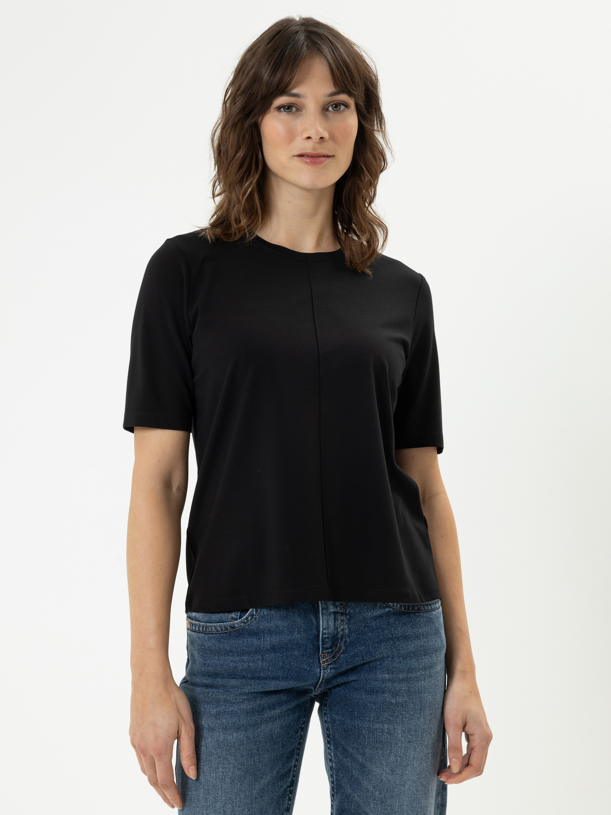 Eine Frau mit gewelltem braunem Haar trägt ein schwarzes kurzärmeliges Oberteil und eine blaue Jeans. Sie steht vor einem schlichten weißen Hintergrund und schaut mit neutralem Blick in die Kamera.