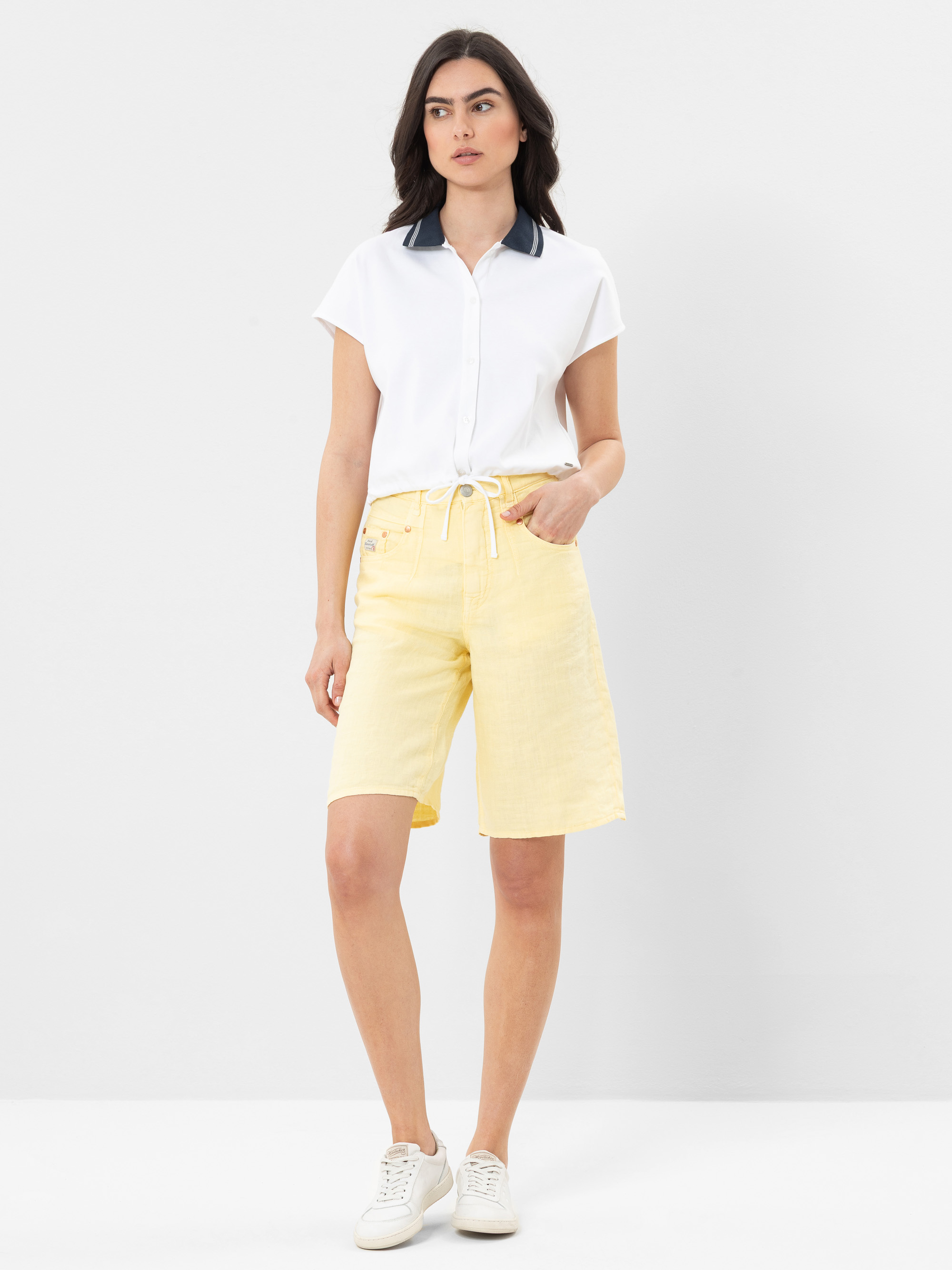 Eine Frau steht vor einem schlichten, hellen Hintergrund und trägt eine kurzärmelige weiße Bluse mit dunklem Kragen, hellgelbe Shorts mit Kordelzug und weiße Turnschuhe. Sie hat dunkles Haar und einen neutralen Gesichtsausdruck.