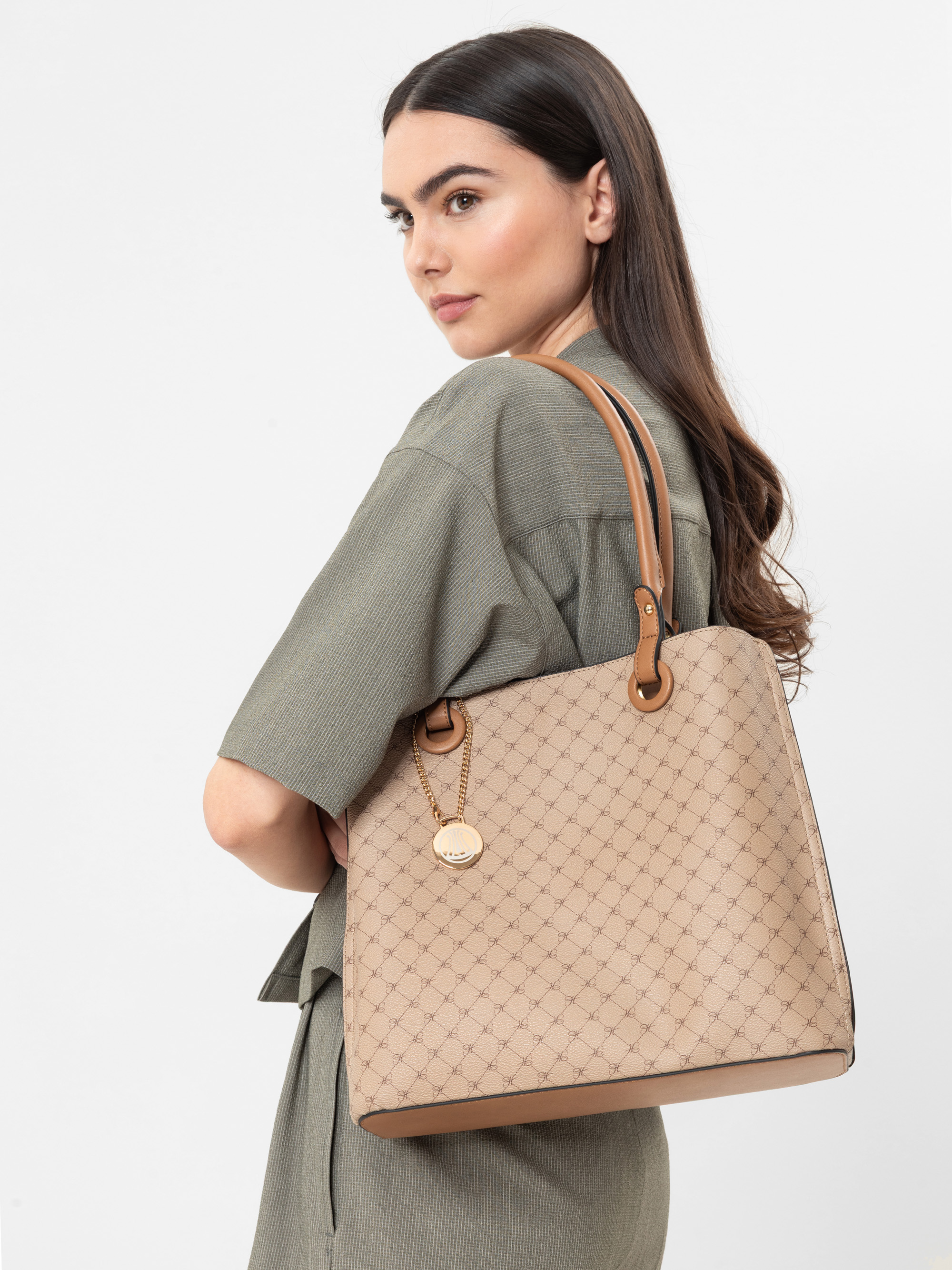 Eine Frau mit langen braunen Haaren trägt ein grünes Outfit und hat eine beige gemusterte Handtasche mit braunen Griffen über der Schulter. Sie blickt über ihre Schulter in Richtung der Kamera vor einem schlichten weißen Hintergrund.