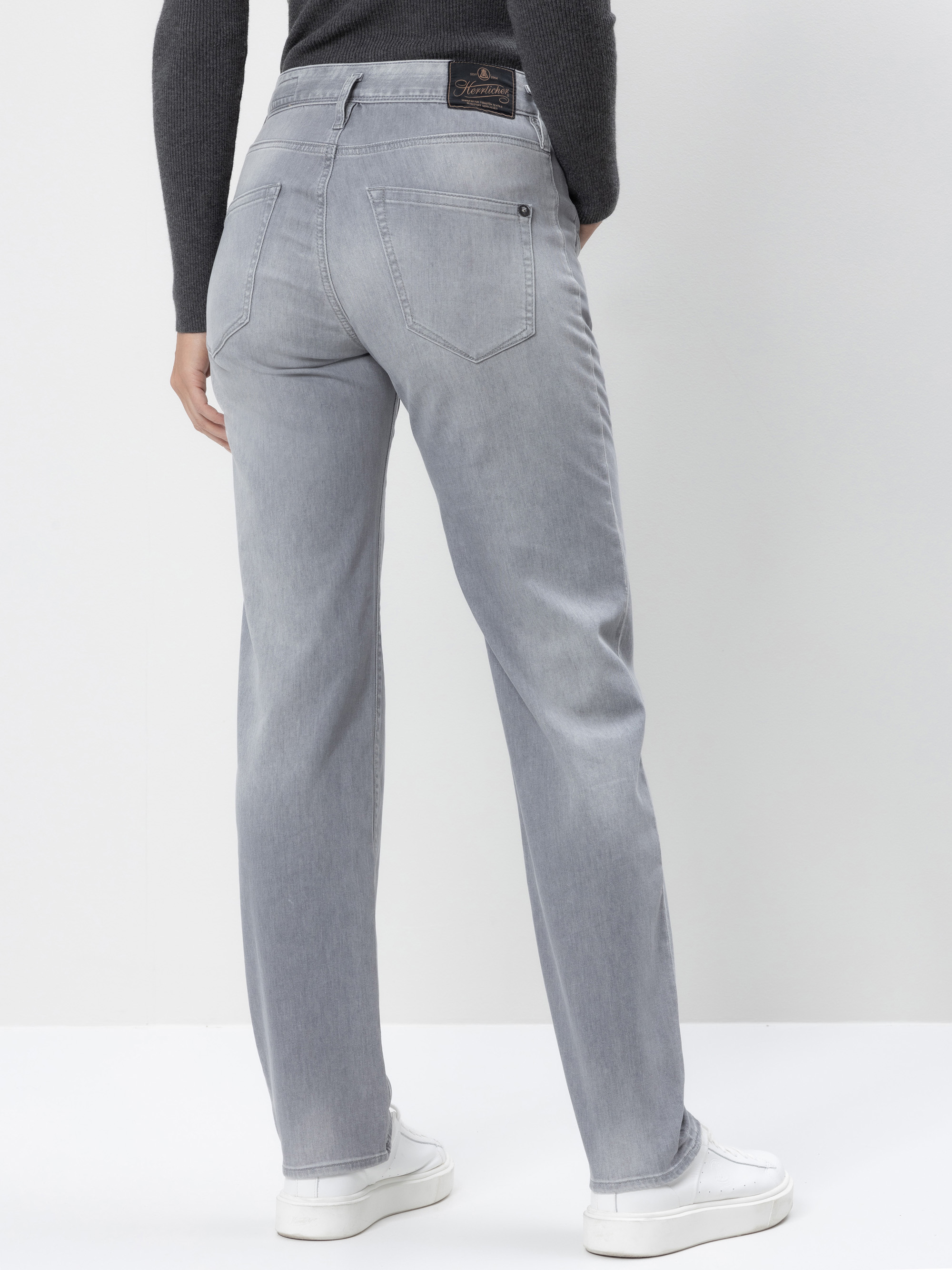 Eine Person, die hellgraue Jeans mit geradem Bein, ein dunkles langärmeliges Oberteil und weiße Turnschuhe trägt, steht mit dem Rücken zur Kamera vor einer weißen Wand.