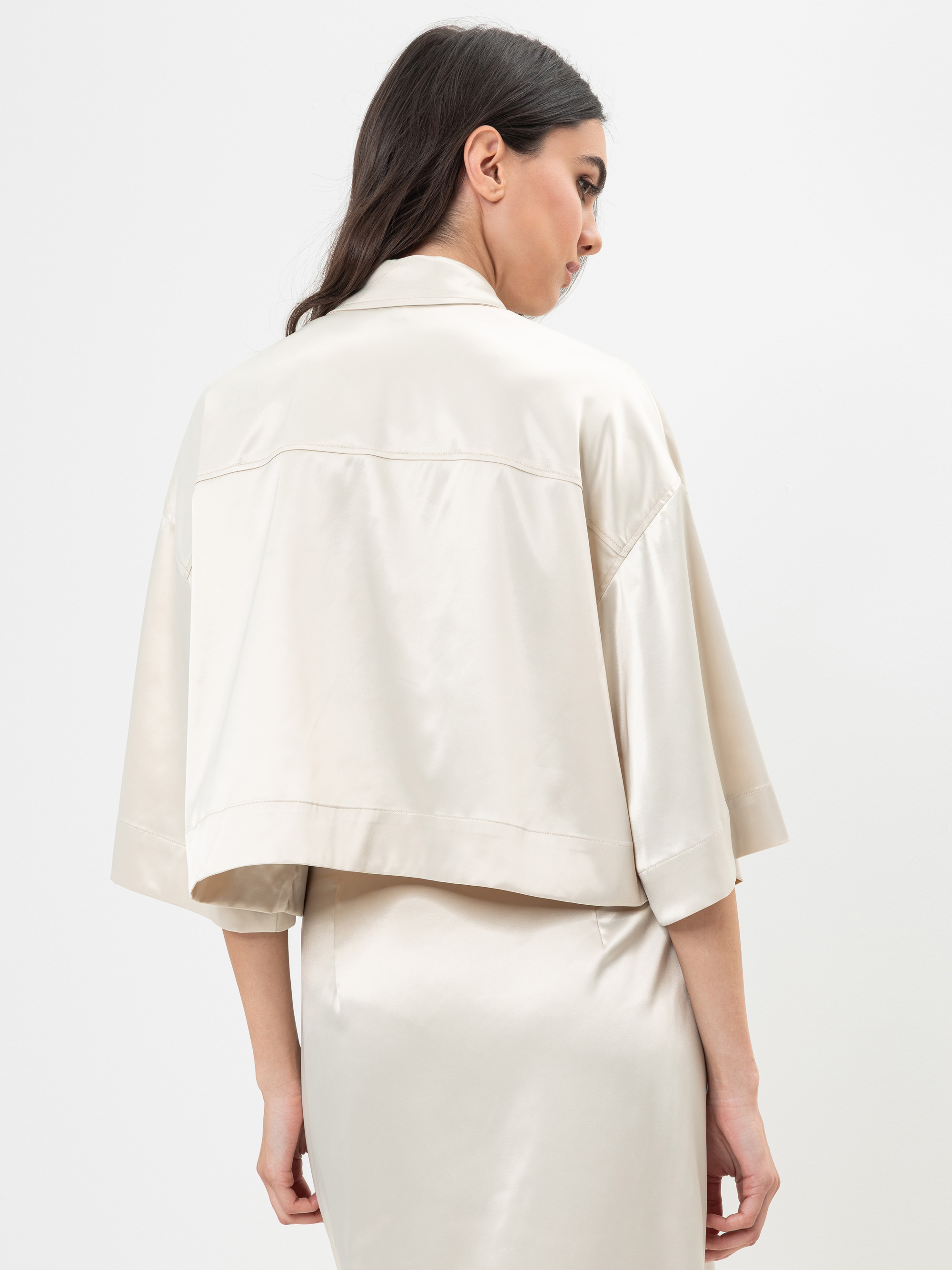 Eine Frau mit langen dunklen Haaren trägt eine cremefarbene, glänzende, kurz geschnittene Jacke mit weiten Ärmeln und einen dazu passenden Rock und steht mit dem Rücken zur Kamera vor einem schlichten weißen Hintergrund.