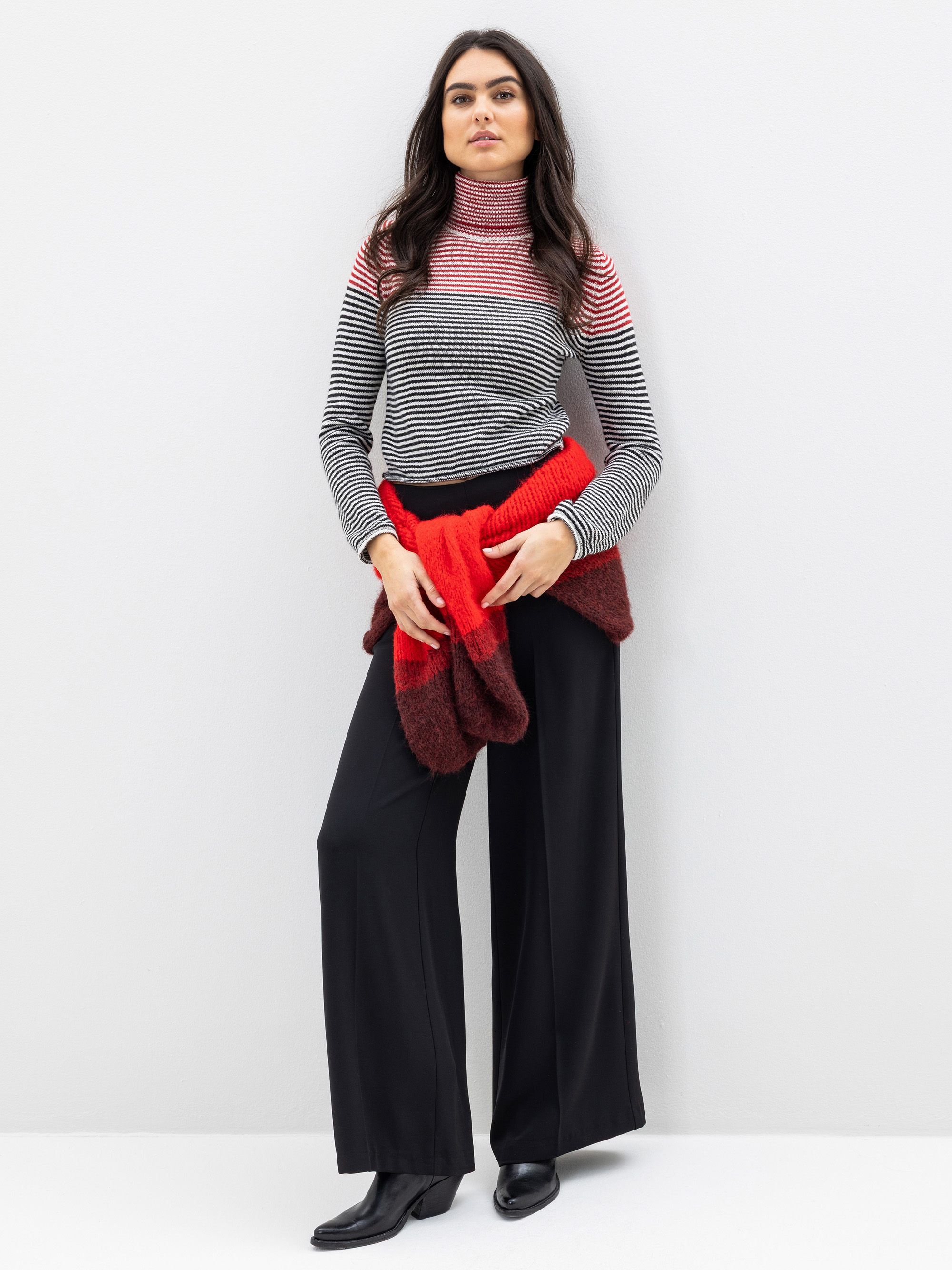 Eine Frau steht vor einer weißen Wand und trägt einen gestreiften Rollkragenpullover, eine schwarze Hose mit weitem Bein, schwarze Stiefel und einen um die Taille gebundenen roten Pullover. Sie hat langes dunkles Haar und einen neutralen Gesichtsausdruc