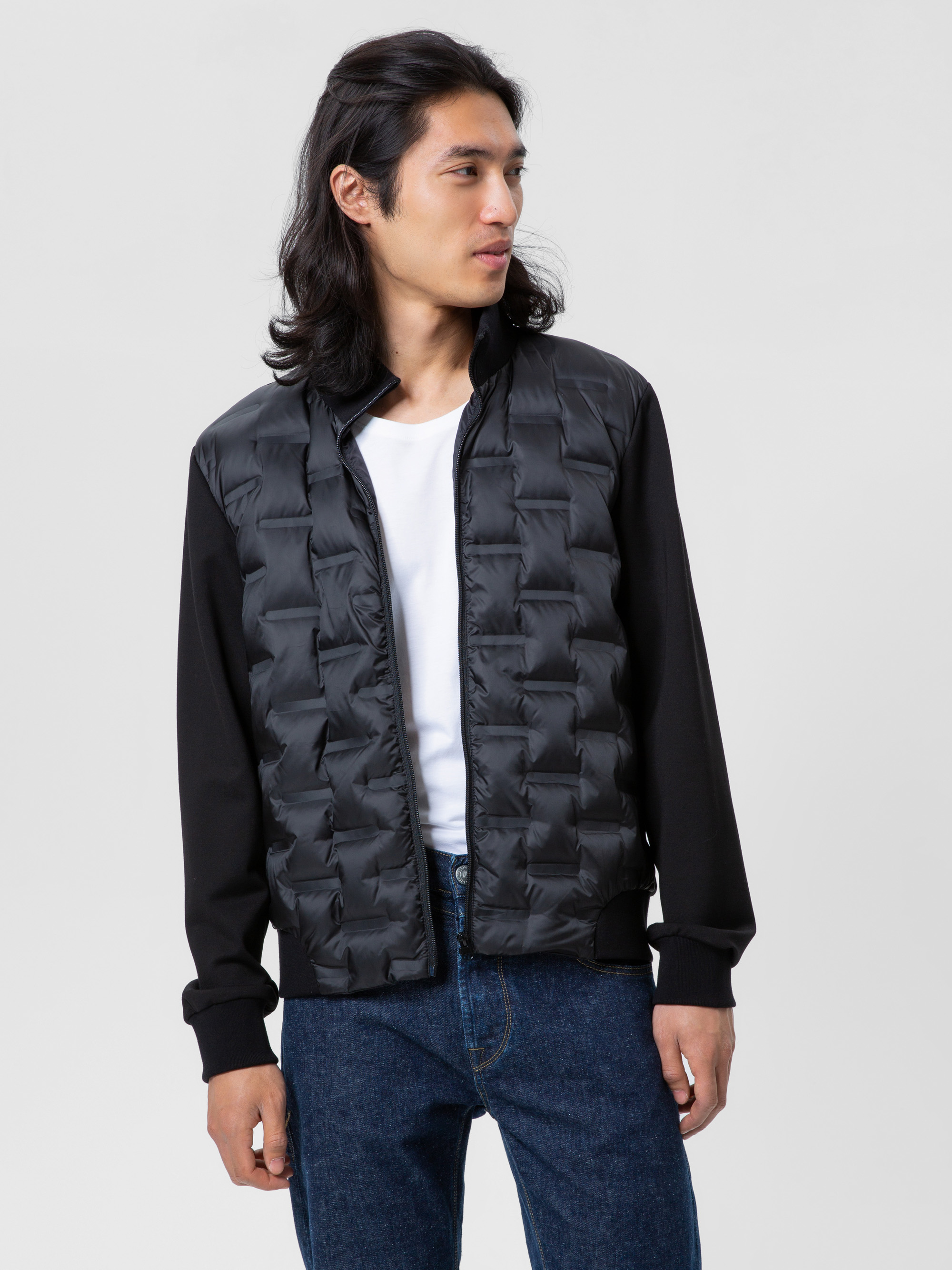 Eine Person mit langen dunklen Haaren trägt eine schwarze Steppjacke über einem weißen T-Shirt und blaue Jeans, steht vor einem schlichten hellen Hintergrund und blickt zur Seite.