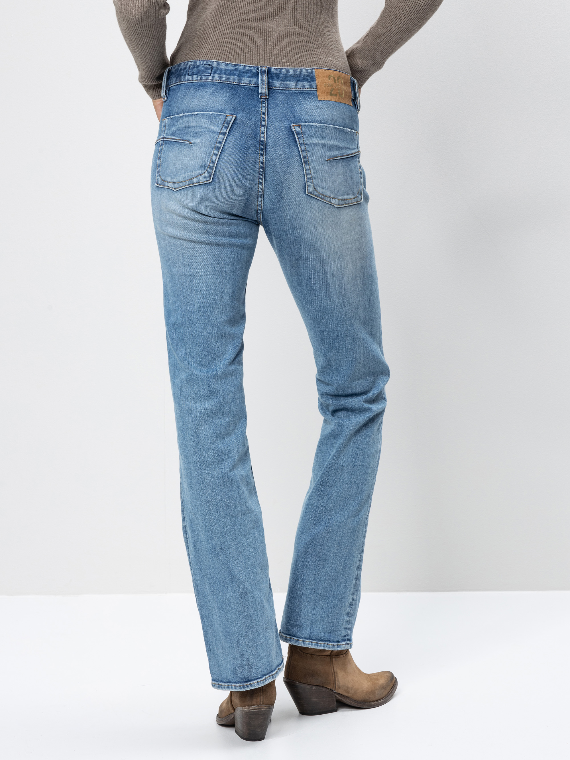 Eine Person in hellblauen Jeans mit geradem Bein, braunen Stiefeln und einem taupefarbenen Pullover steht vor einem schlichten weißen Hintergrund, von hinten gesehen.