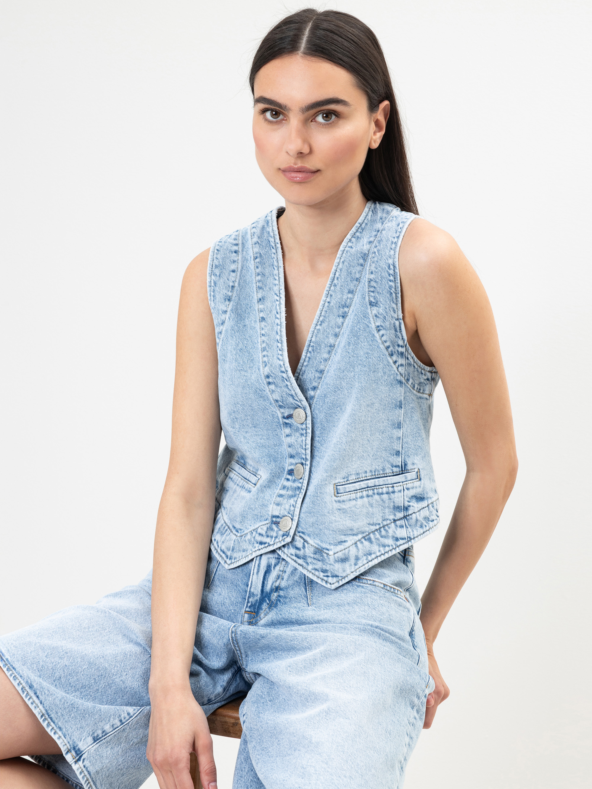 Eine Frau mit langen dunklen Haaren trägt eine hellblaue ärmellose Jeansweste und dazu passende Jeans-Shorts mit hoher Taille, die vor einem schlichten weißen Hintergrund stehen.
