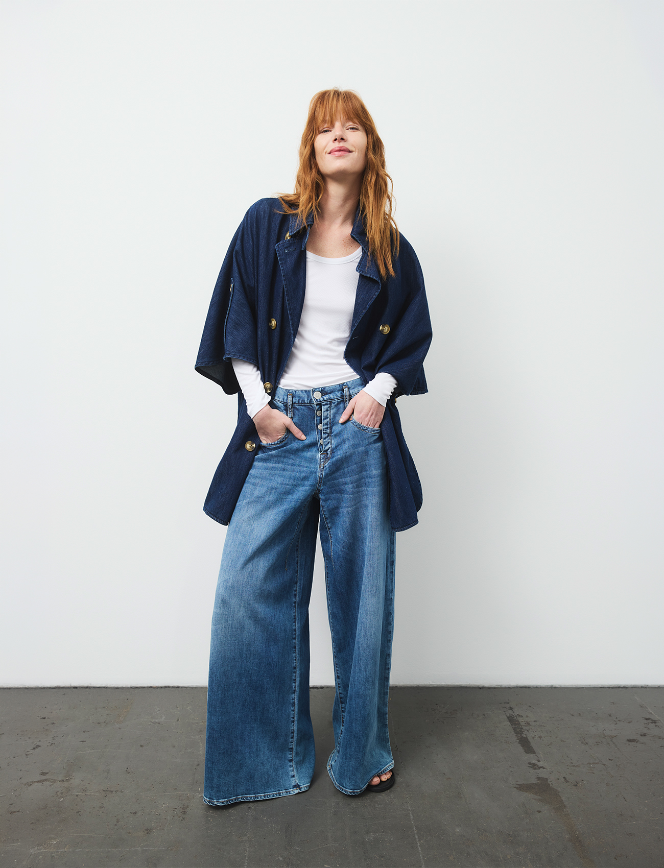 Eine Frau mit langen roten Haaren trägt eine marineblaue Jeansjacke über einem weißen Oberteil und einer blau, weit geschnittenen Jeans. Sie steht vor einer schlichten weißen Wand und einem grauen Boden, lächelt und hat die Hände in den Taschen.