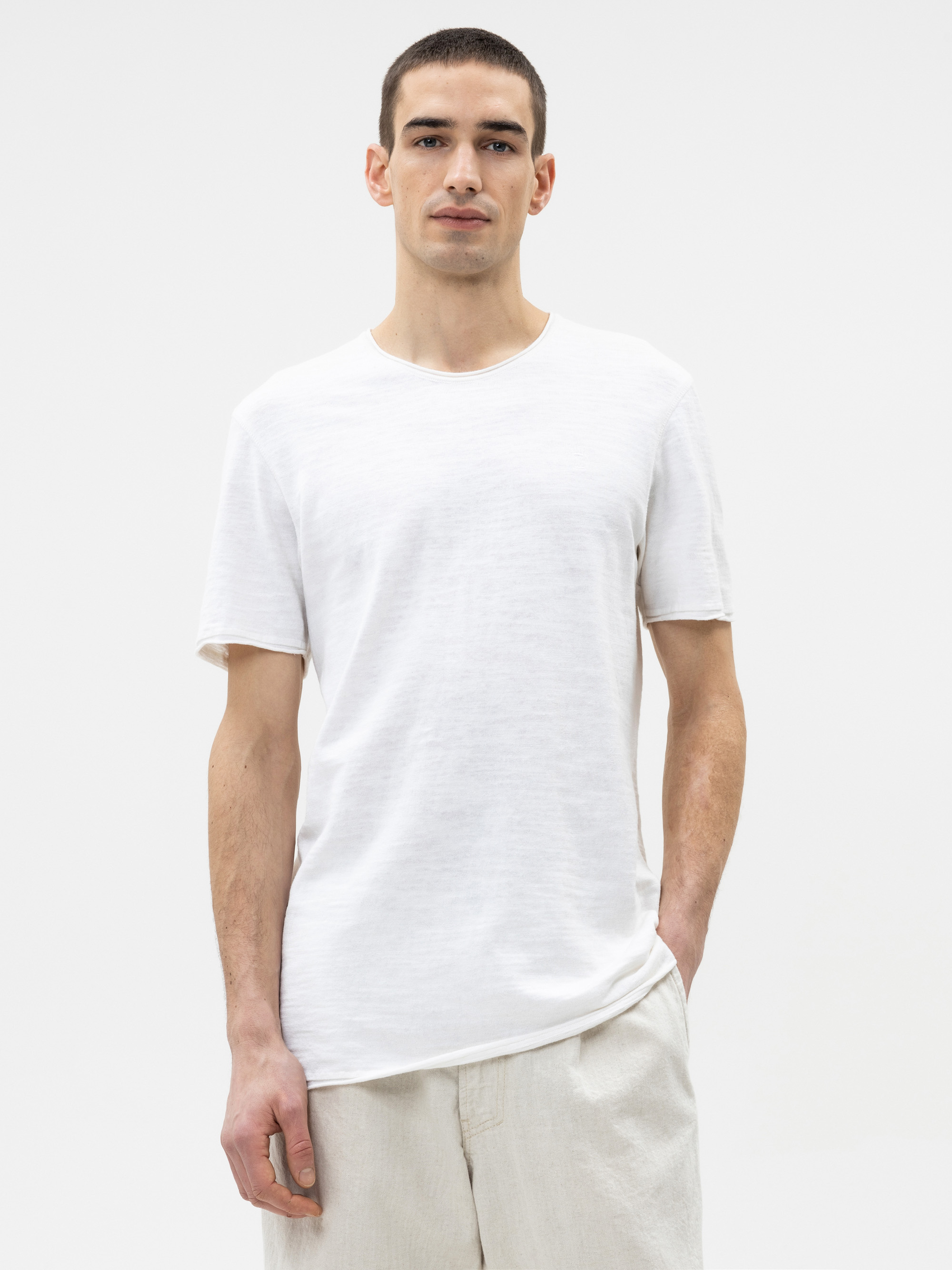 Ein Mann, der ein schlichtes weißes, kurzärmeliges T-Shirt und eine hellbeige Hose trägt, steht vor einem weißen Hintergrund und schaut mit neutralem Blick direkt in die Kamera.