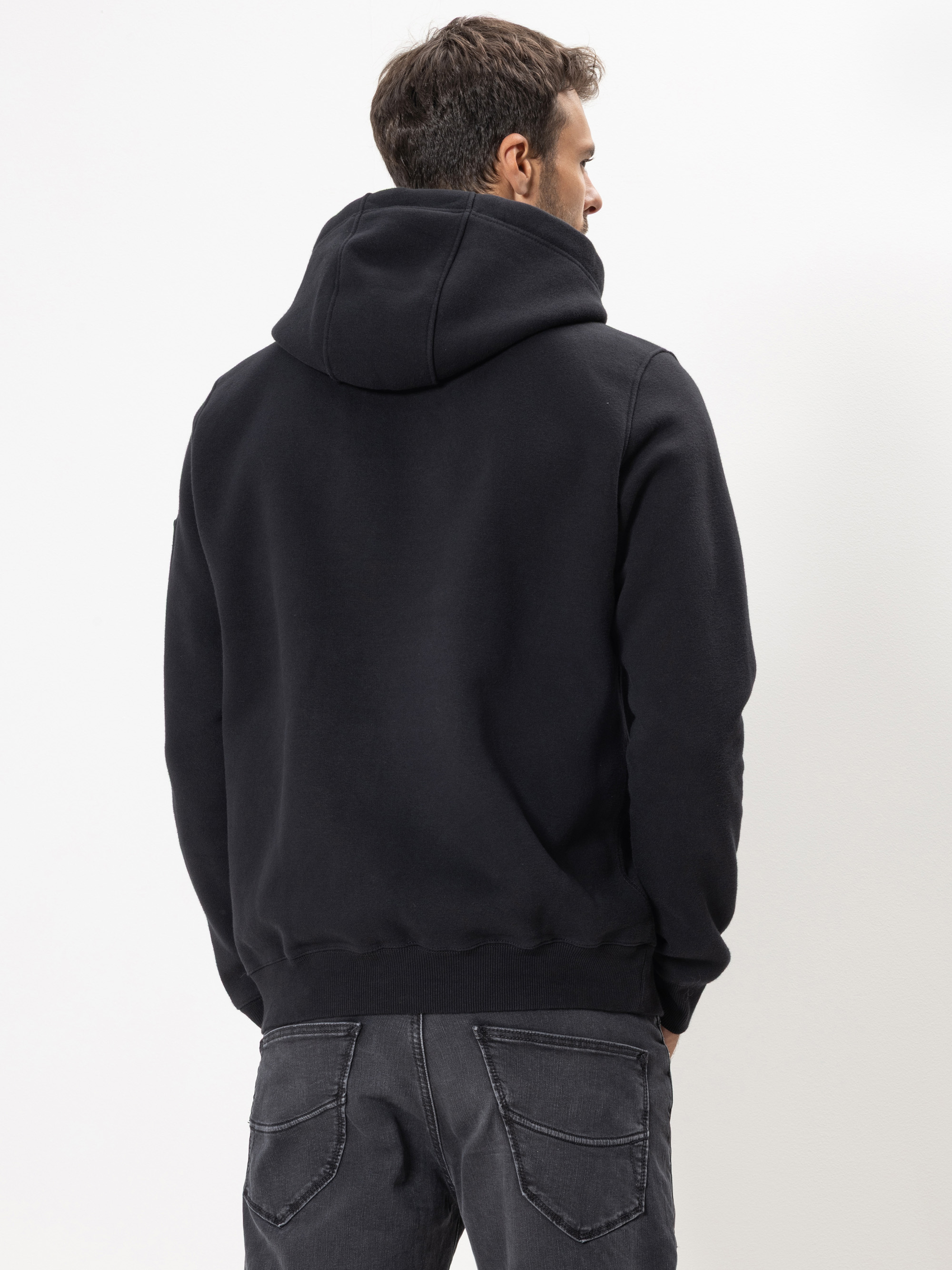 Ein Mann steht mit dem Rücken zur Kamera, trägt ein schwarzes Sweatshirt mit Kapuze und dunkelgraue Jeans vor einem schlichten weißen Hintergrund.