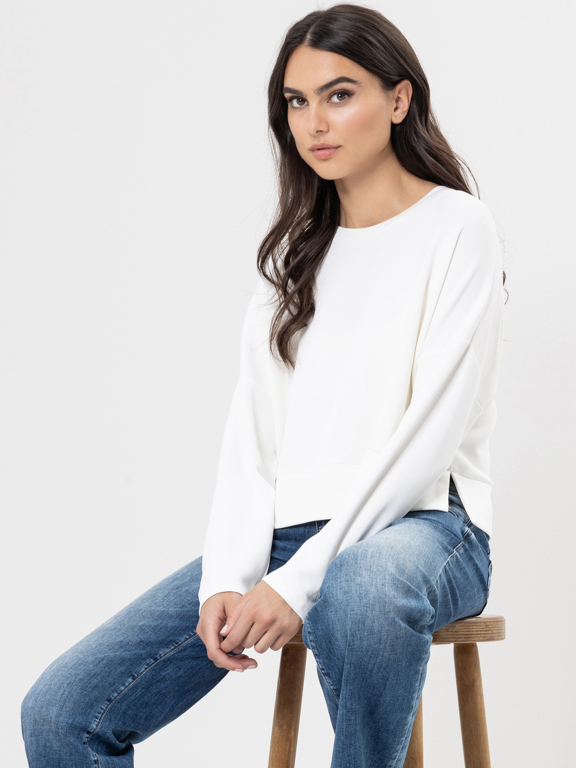 Eine Frau mit langen dunklen Haaren sitzt auf einem Holzschemel, trägt einen weiten weißen Pullover und eine blaue Jeans und blickt leicht zur Seite mit einem neutralen Ausdruck vor einem schlichten hellen Hintergrund.