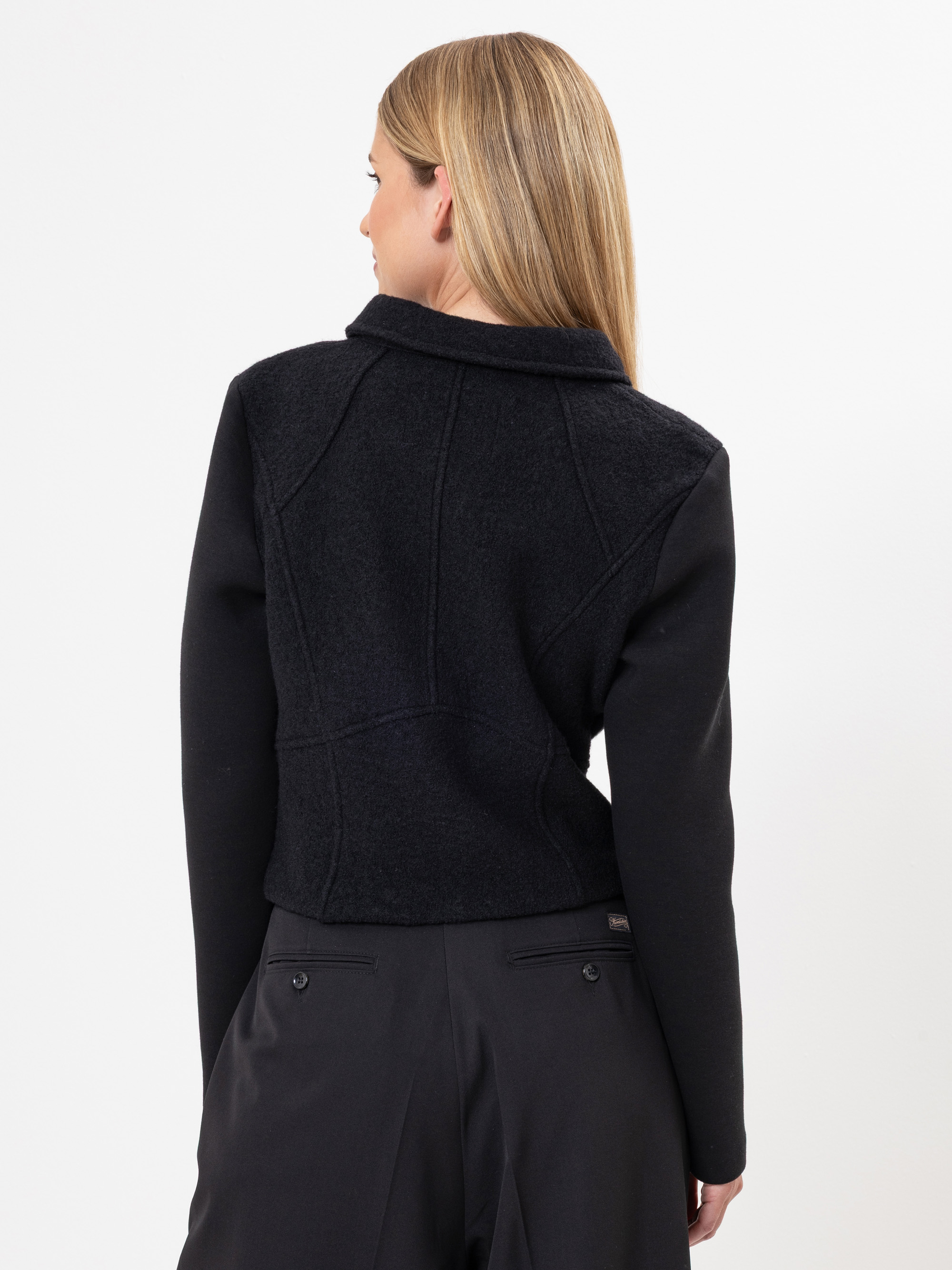 Eine Frau mit glattem, blondem Haar wird von hinten gezeigt. Sie trägt eine schwarze, maßgeschneiderte Jacke und eine schwarze Hose vor einem schlichten weißen Hintergrund.