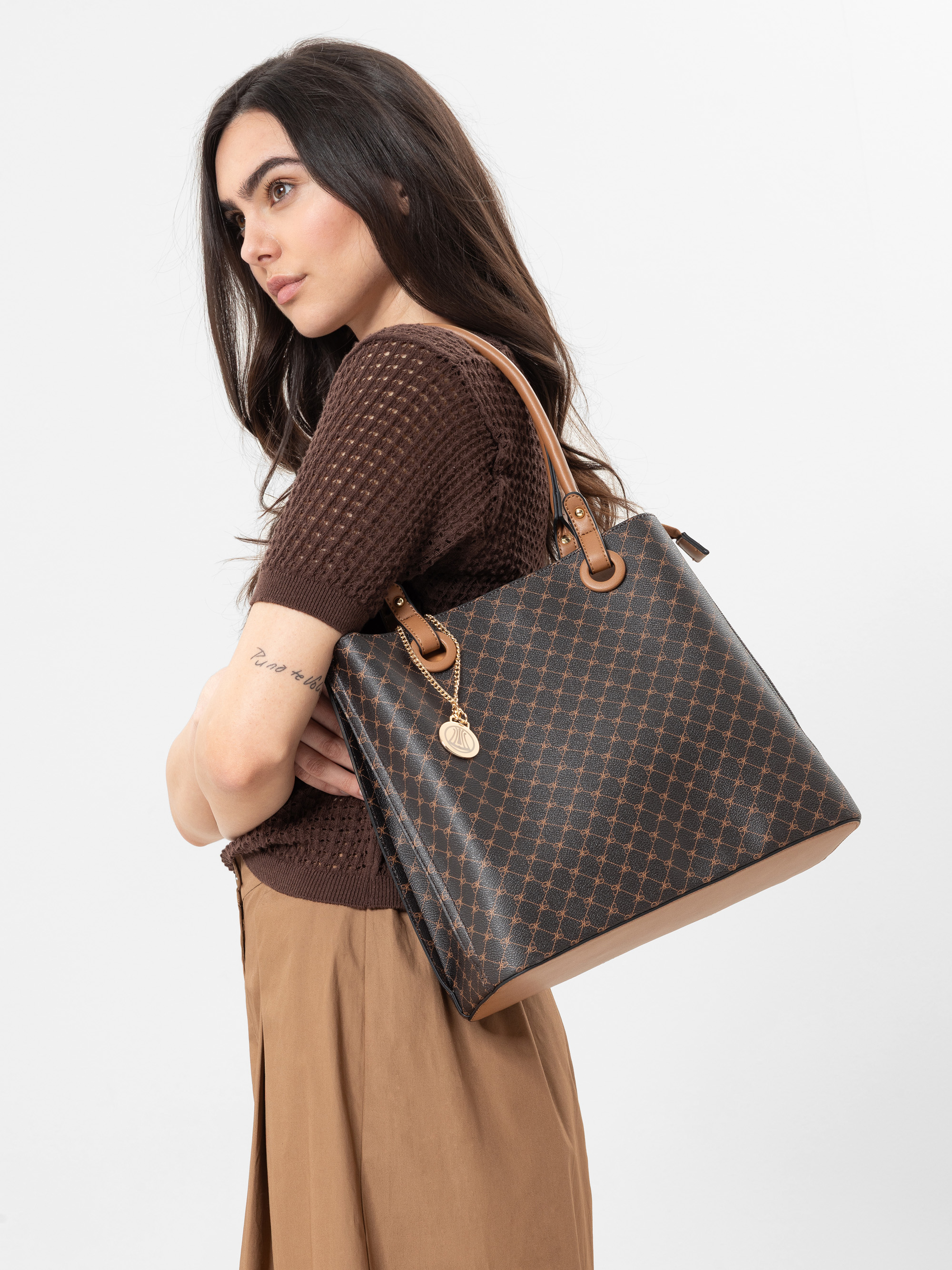 Eine Frau mit langen dunklen Haaren trägt ein braunes Strickoberteil und einen hellbraunen Rock und trägt eine große braun gemusterte Tragetasche mit hellbraunen Trägern über der Schulter. Sie steht vor einem schlichten weißen Hintergrund.