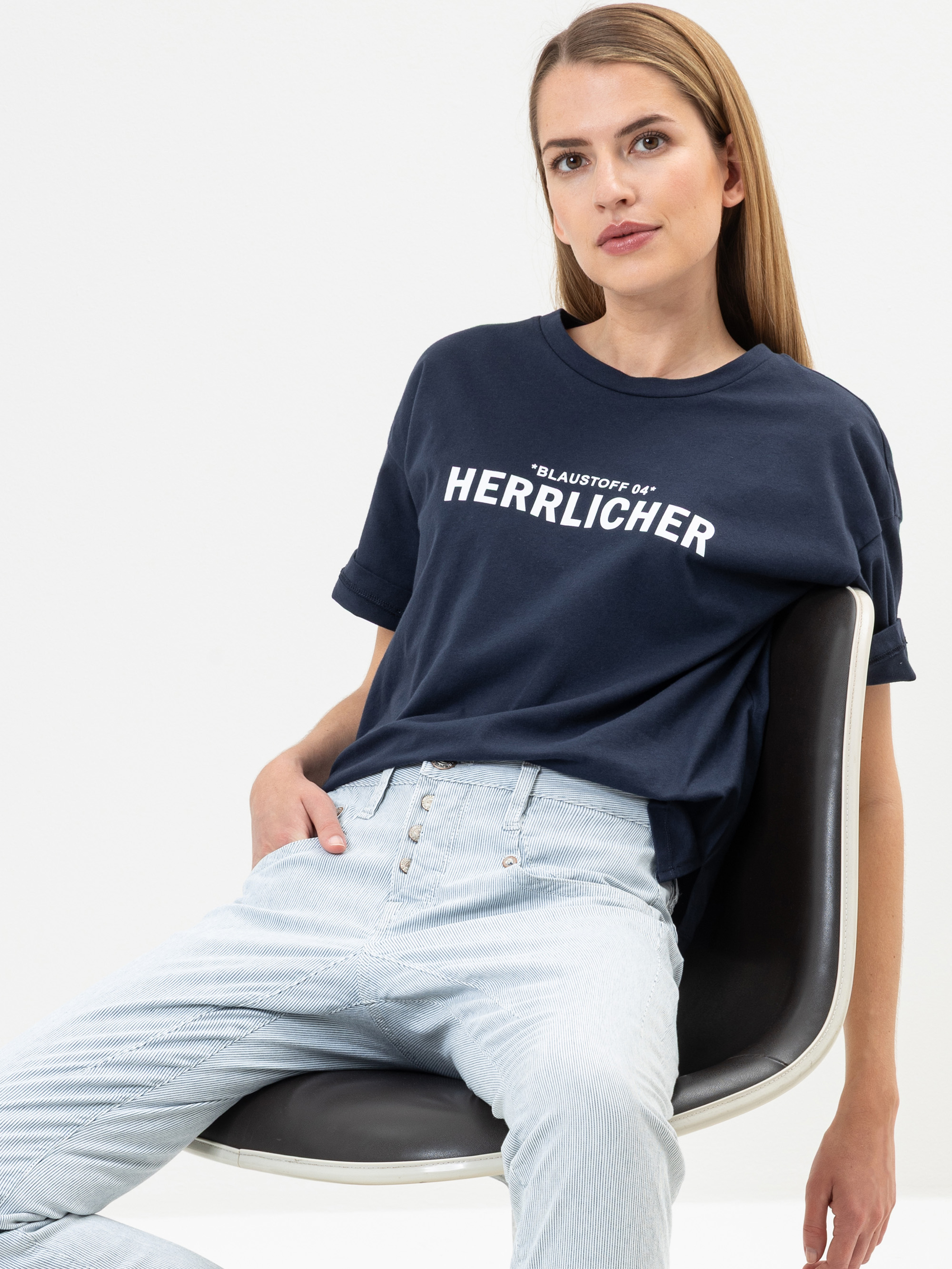 Eine Frau mit glattem, hellbraunem Haar sitzt auf einem modernen Stuhl und trägt ein marineblaues T-Shirt mit der weißen Aufschrift BLAUSTOFF 04 HERRLICHER und hellblaue Jeans vor einem schlichten weißen Hintergrund.