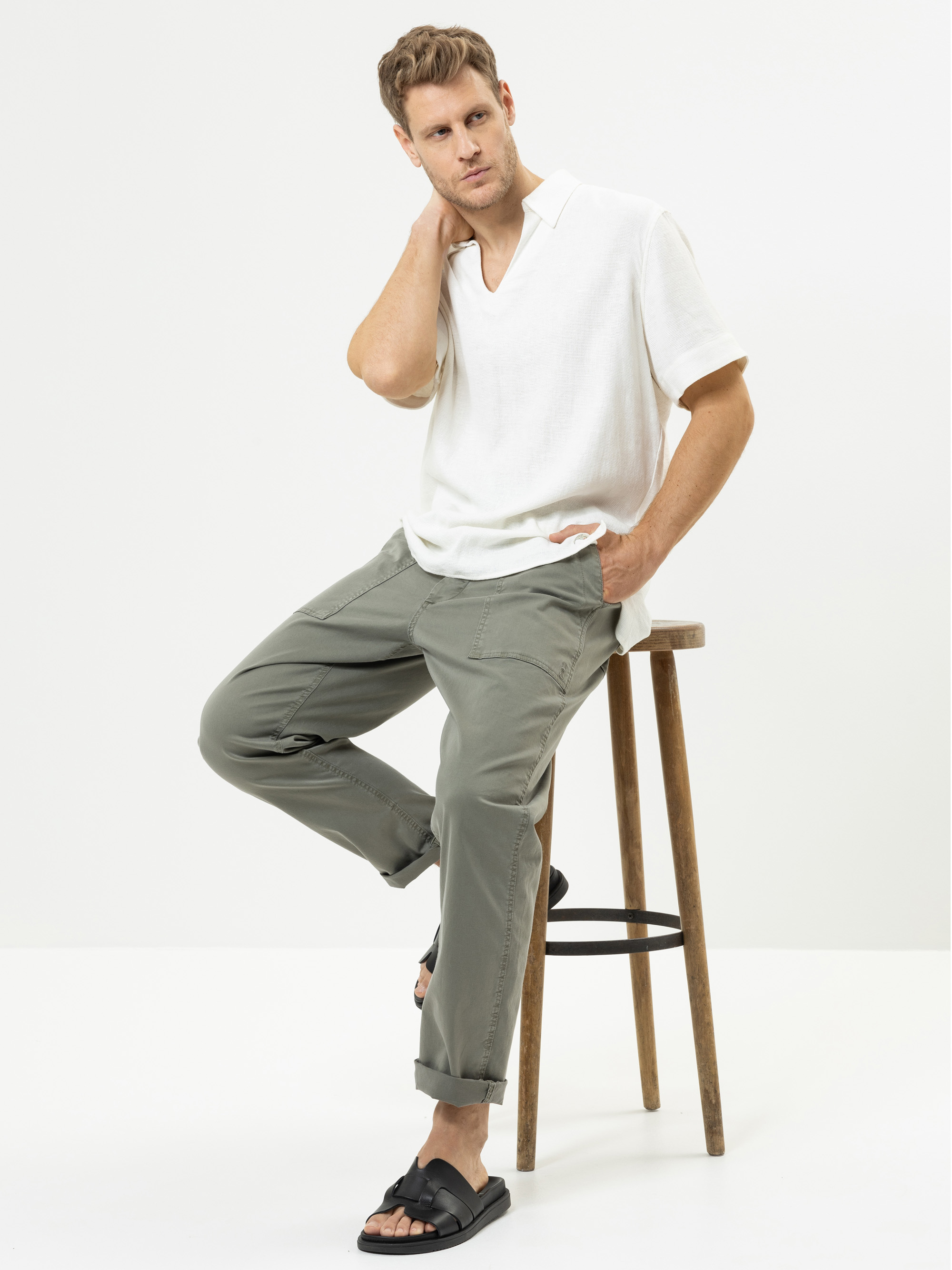 Ein Mann, der ein weißes, kurzärmeliges Hemd, eine olivgrüne Hose und schwarze Sandalen trägt, sitzt auf einem Holzschemel vor einem schlichten weißen Hintergrund und stützt eine Hand in den Nacken, die andere auf den Schemel.