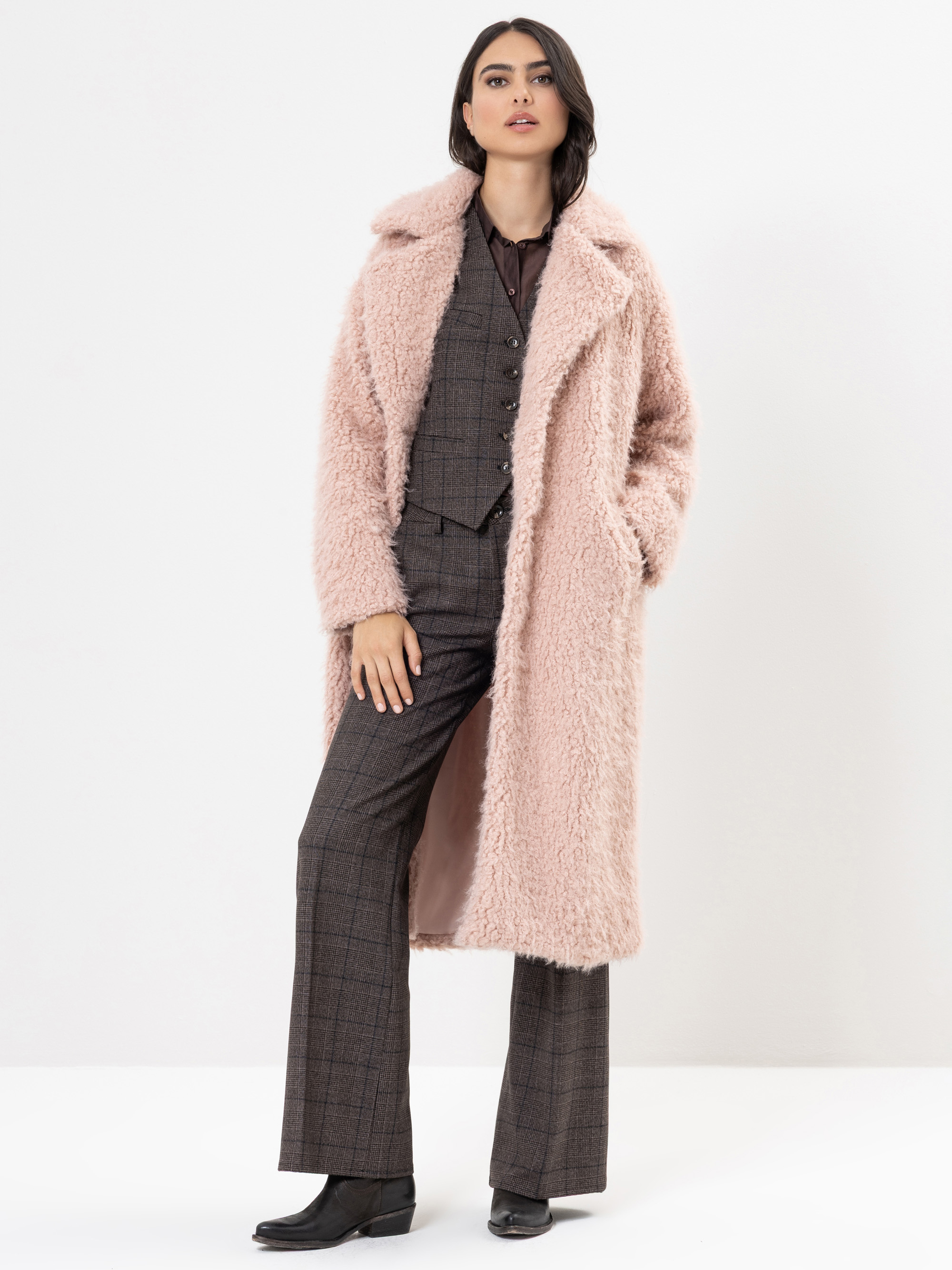Eine Frau steht vor einem weißen Hintergrund und trägt einen strukturierten, hellrosa Kunstpelzmantel über einem braunen, karierten Anzug mit geknöpfter Weste und Schlaghosen, gepaart mit schwarzen Stiefeln. Sie hat dunkles Haar und einen selbstbewuss