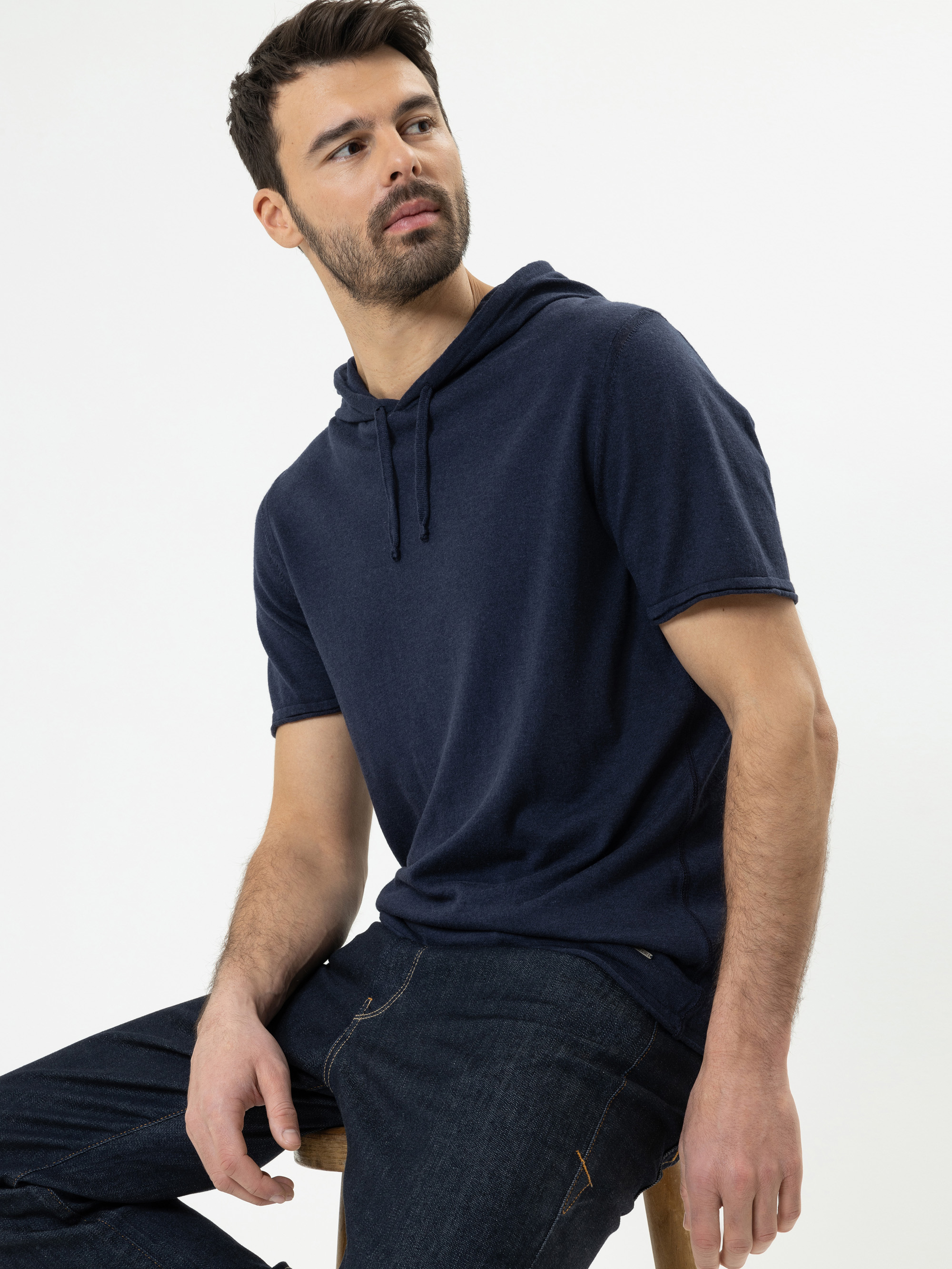 Ein Mann mit kurzen dunklen Haaren und einem Bart sitzt auf einem Hocker, trägt ein marineblaues, kurzärmeliges Kapuzenshirt und dunkle Jeans und blickt leicht zur Seite vor einem schlichten weißen Hintergrund.
