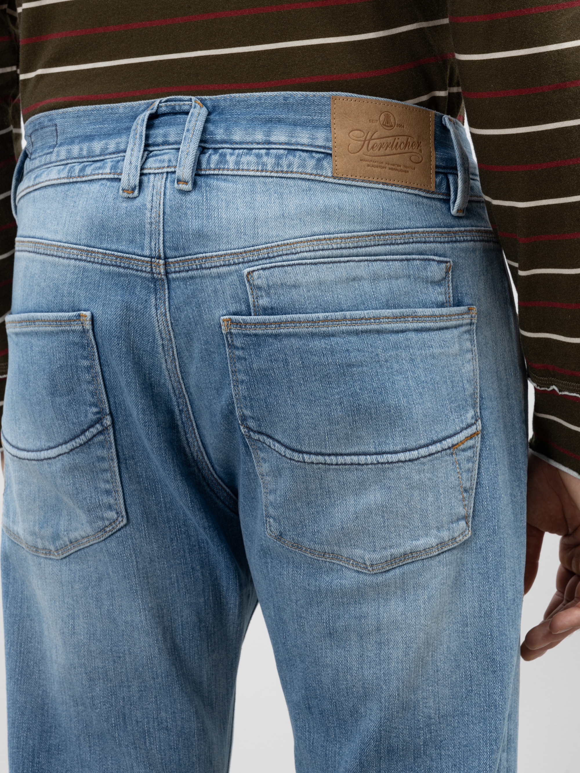 Nahaufnahme einer Person, die eine hellblaue Jeans mit sichtbaren Gesäßtaschen und einem braunen Lederetikett am Bund trägt. Die Person trägt außerdem ein grün und rot gestreiftes Hemd.