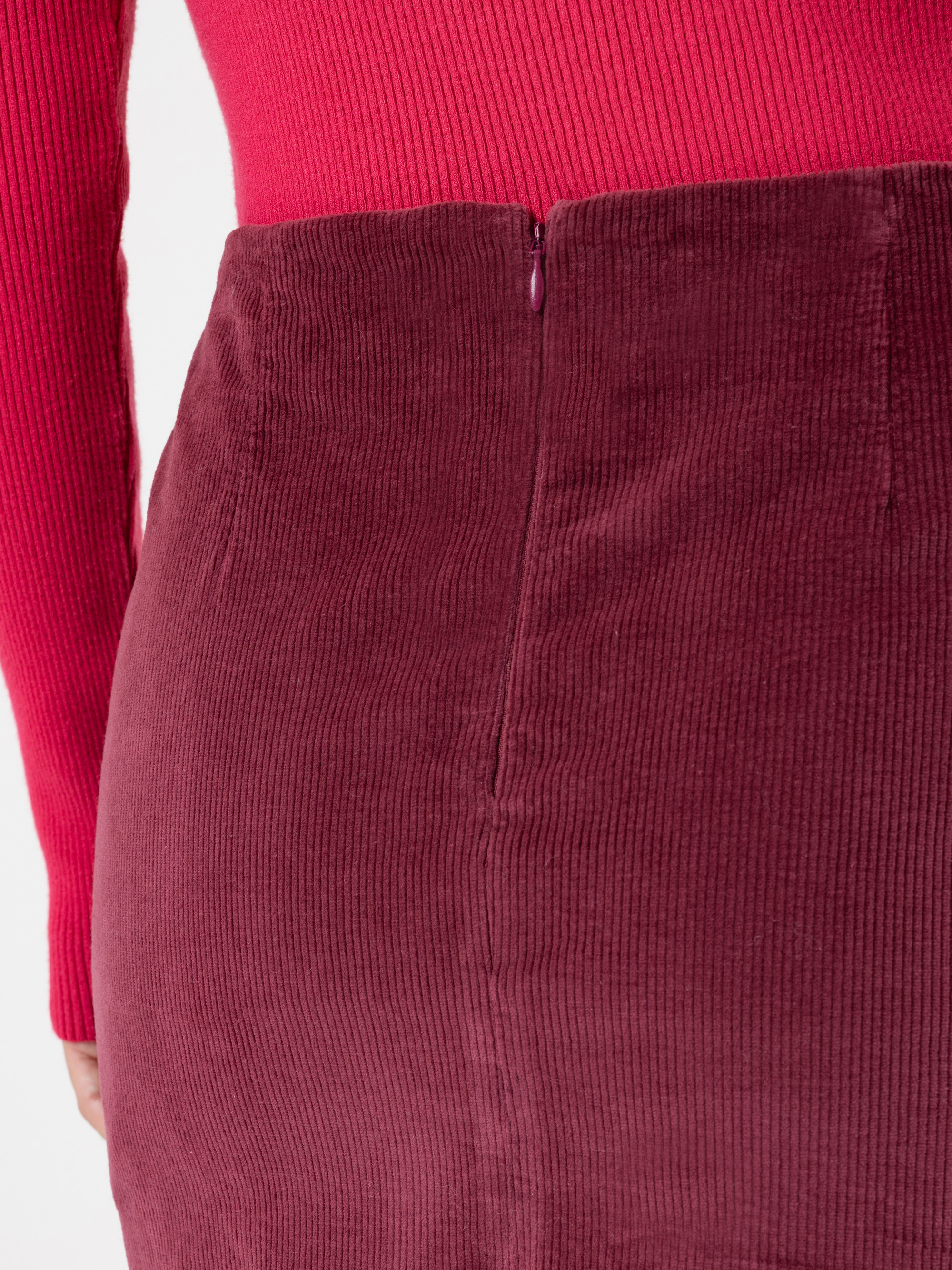 Nahaufnahme einer Person, die ein geripptes, leuchtend rotes Oberteil trägt, das in einen hochtaillierten, strukturierten weinroten Kordrock mit sichtbarem Reißverschluss auf der Rückseite gesteckt wird.