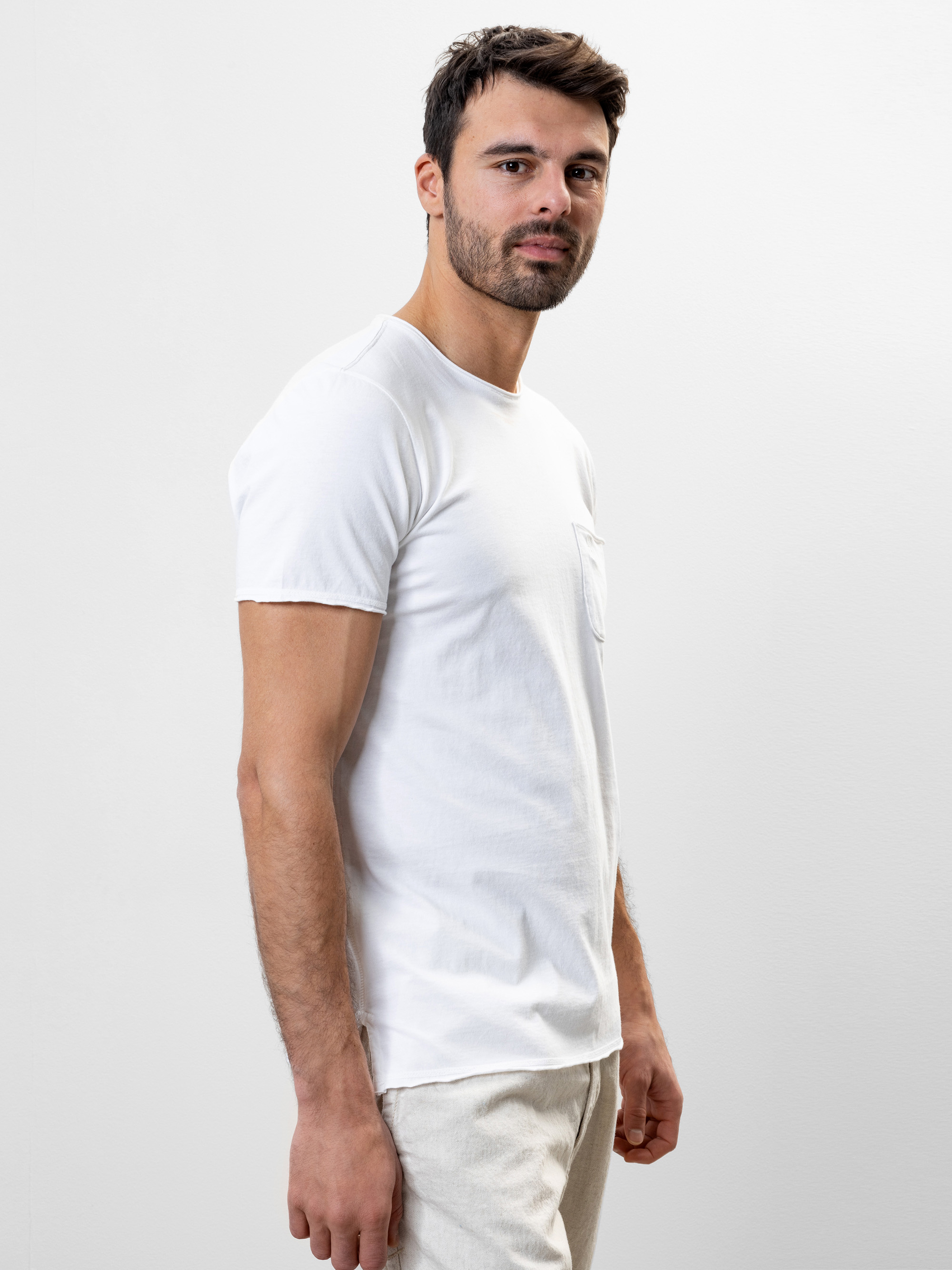 Ein Mann mit kurzen dunklen Haaren und einem Bart steht seitlich, der Kamera zugewandt, in einem einfachen weißen T-Shirt und einer hellen Hose vor einem einfachen weißen Hintergrund.