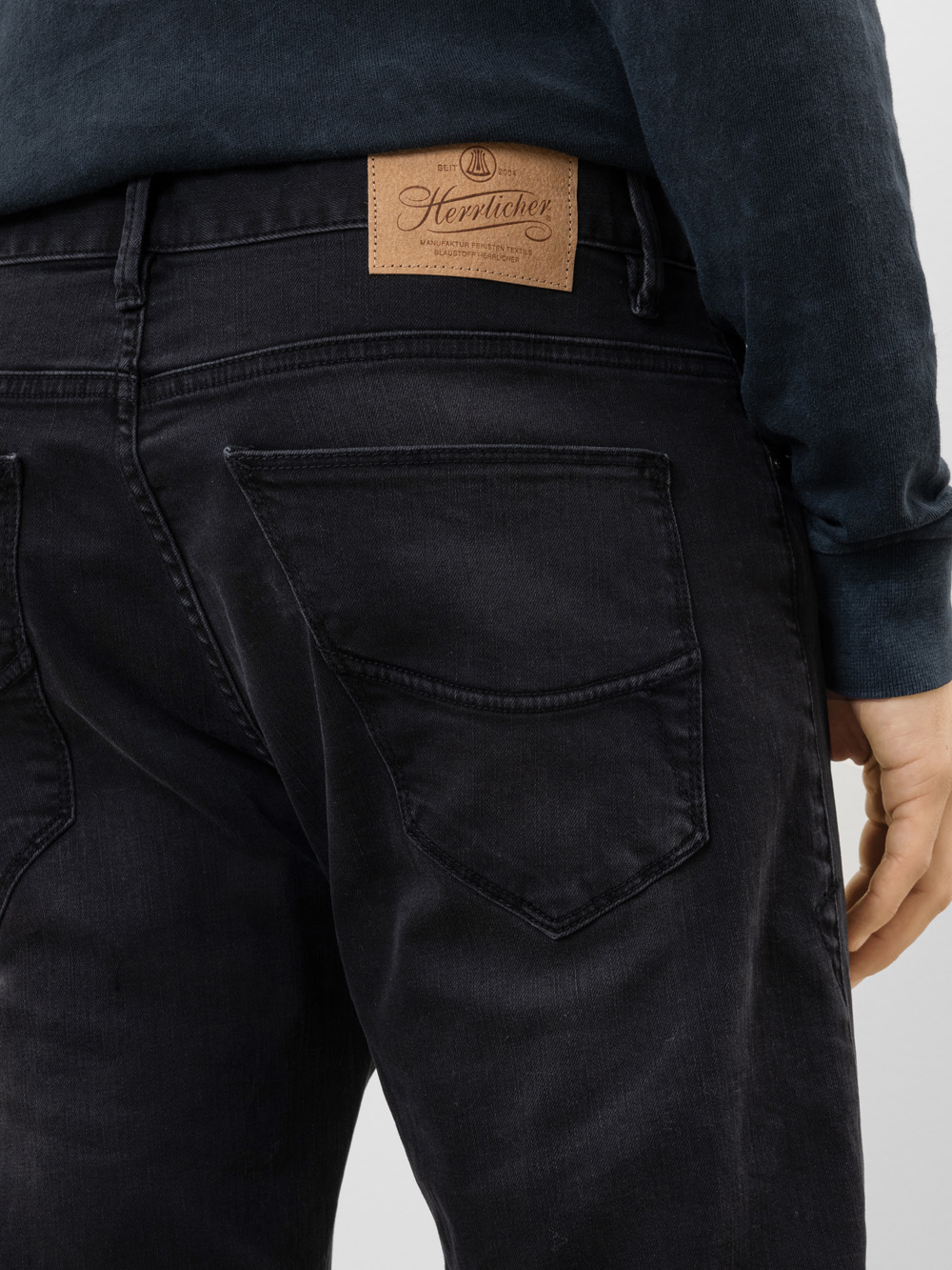 Eine Person trägt eine schwarze Jeans und ein dunkles Oberteil, von hinten gesehen. Die Jeans hat große Gesäßtaschen und einen hellbraunen Lederaufnäher am Bund, auf dem der Markenname Herrlicher zu sehen ist.