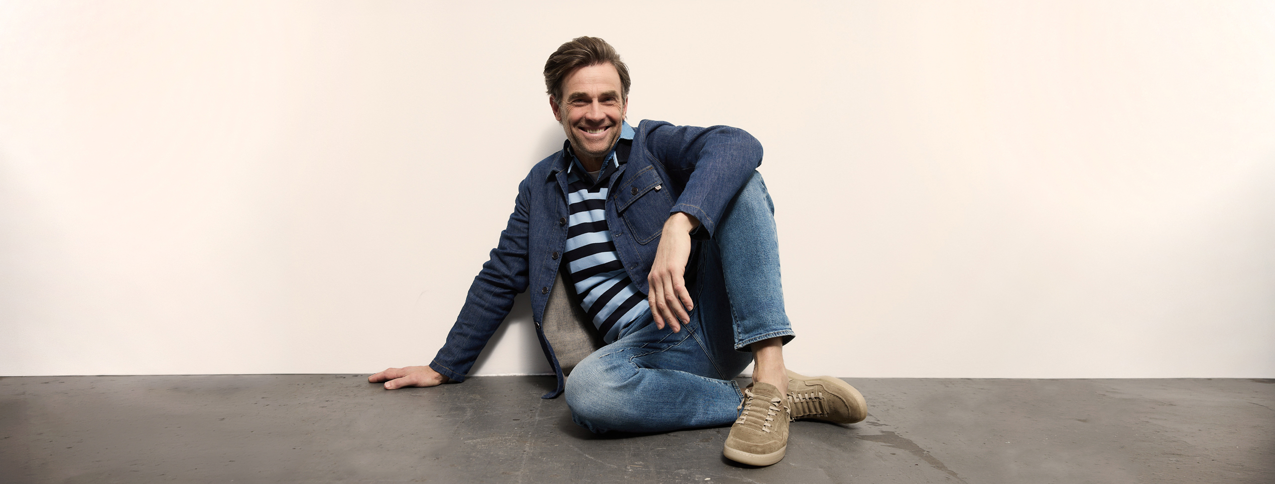 Ein Mann in blauer Jacke, gestreiftem Hemd und Jeans sitzt lässig auf dem Boden vor einer weißen Wand, lächelt, hat ein Bein gekreuzt und einen Arm auf den Boden gestützt.