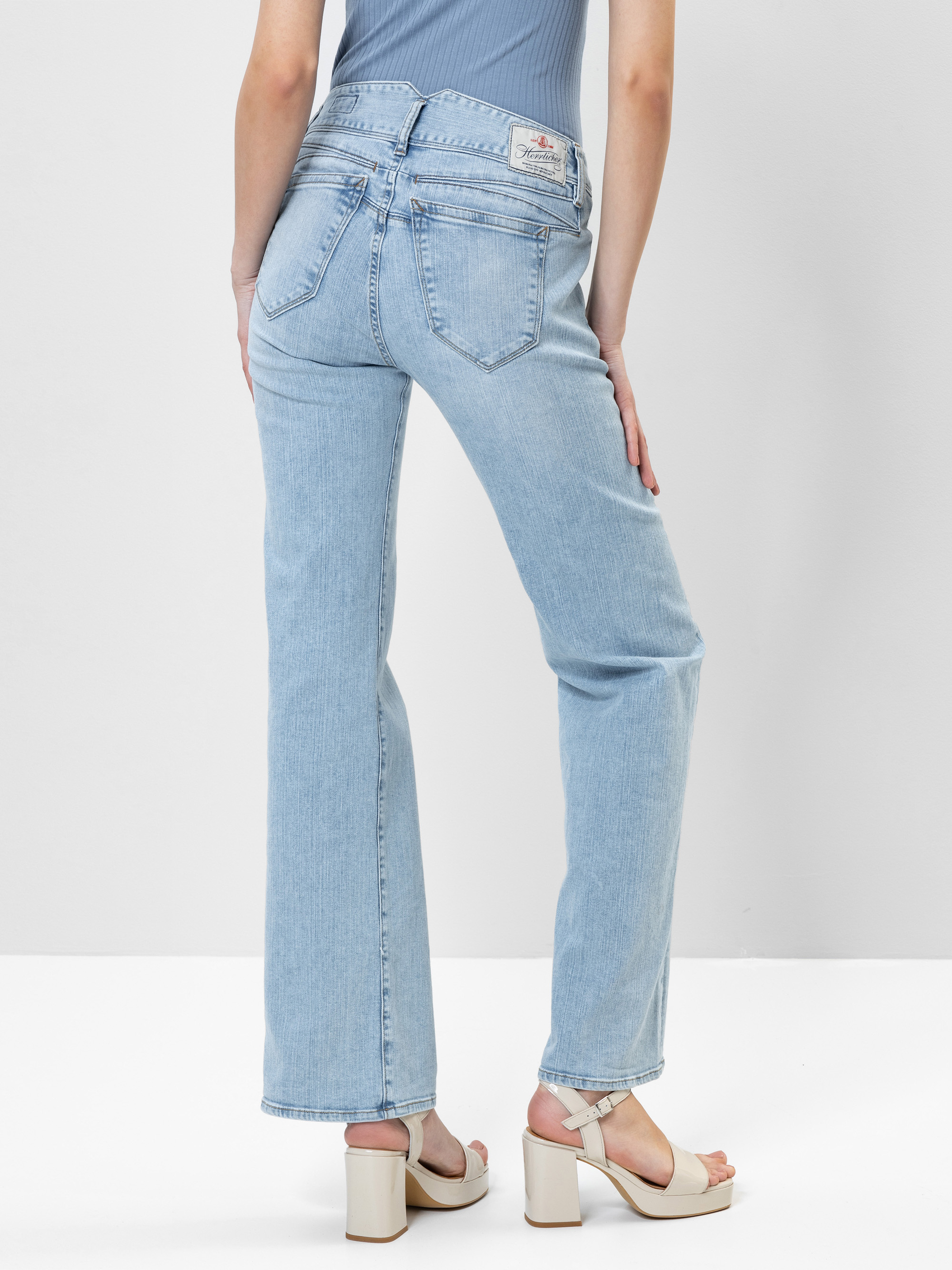 Eine Person, die hellblaue Jeans mit geradem Bein und cremefarbene Plateausandalen trägt, steht mit einer Hand an der Hüfte vor einem schlichten weißen Hintergrund.