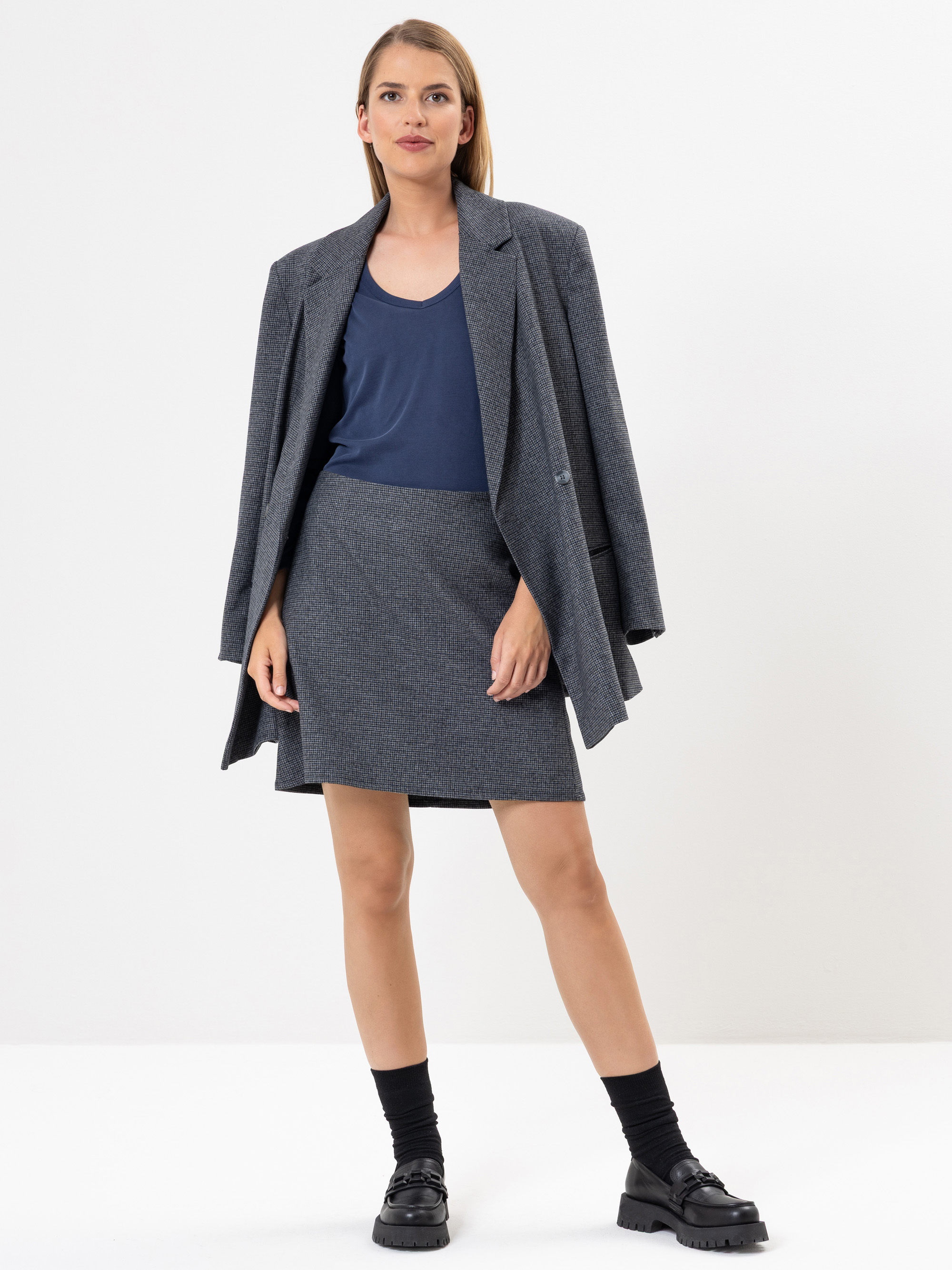 Eine Frau steht in einem grauen Rock und einem passenden Blazer, der über ihre Schultern drapiert ist, mit einem marineblauen Oberteil, schwarzen Socken und schwarzen klobigen Schuhen vor einem schlichten weißen Hintergrund.