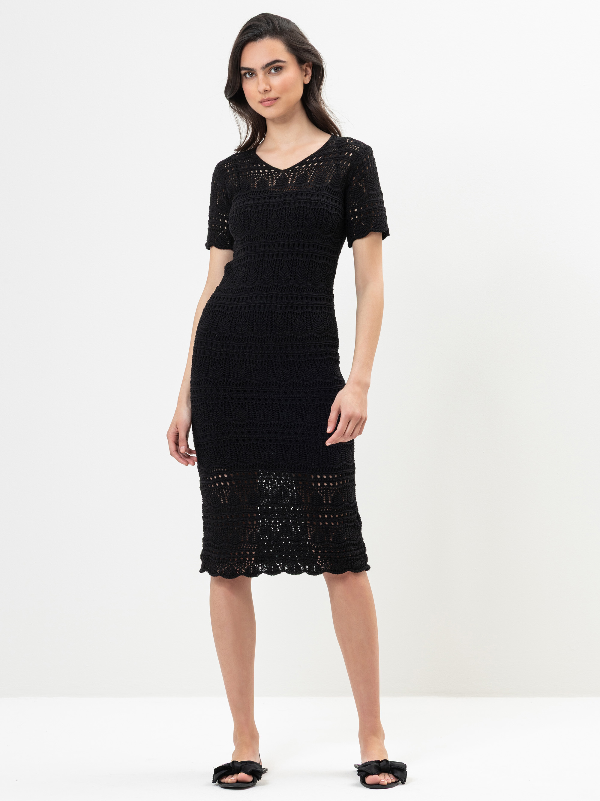 Eine Frau steht vor einem schlichten, hellen Hintergrund. Sie trägt ein kurzärmeliges, schwarzes Häkelkleid, das bis unter das Knie reicht, und schwarze, offene Sandalen mit einer Schleife. Sie hat dunkles Haar und einen neutralen Gesichtsausdruck.