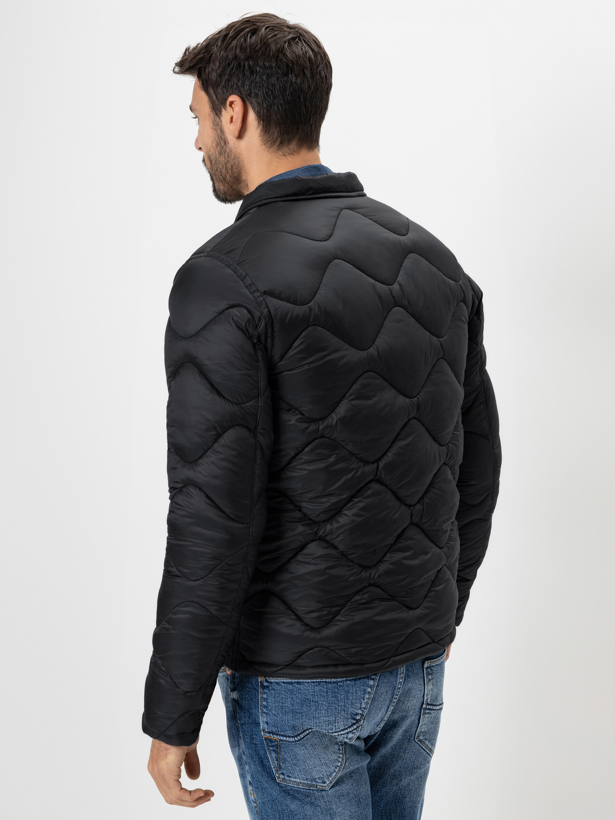 Ein Mann mit dunklem Haar, von hinten gesehen, trägt eine schwarze Steppjacke mit Wellenmuster und blaue Jeans. Er steht vor einem schlichten weißen Hintergrund.