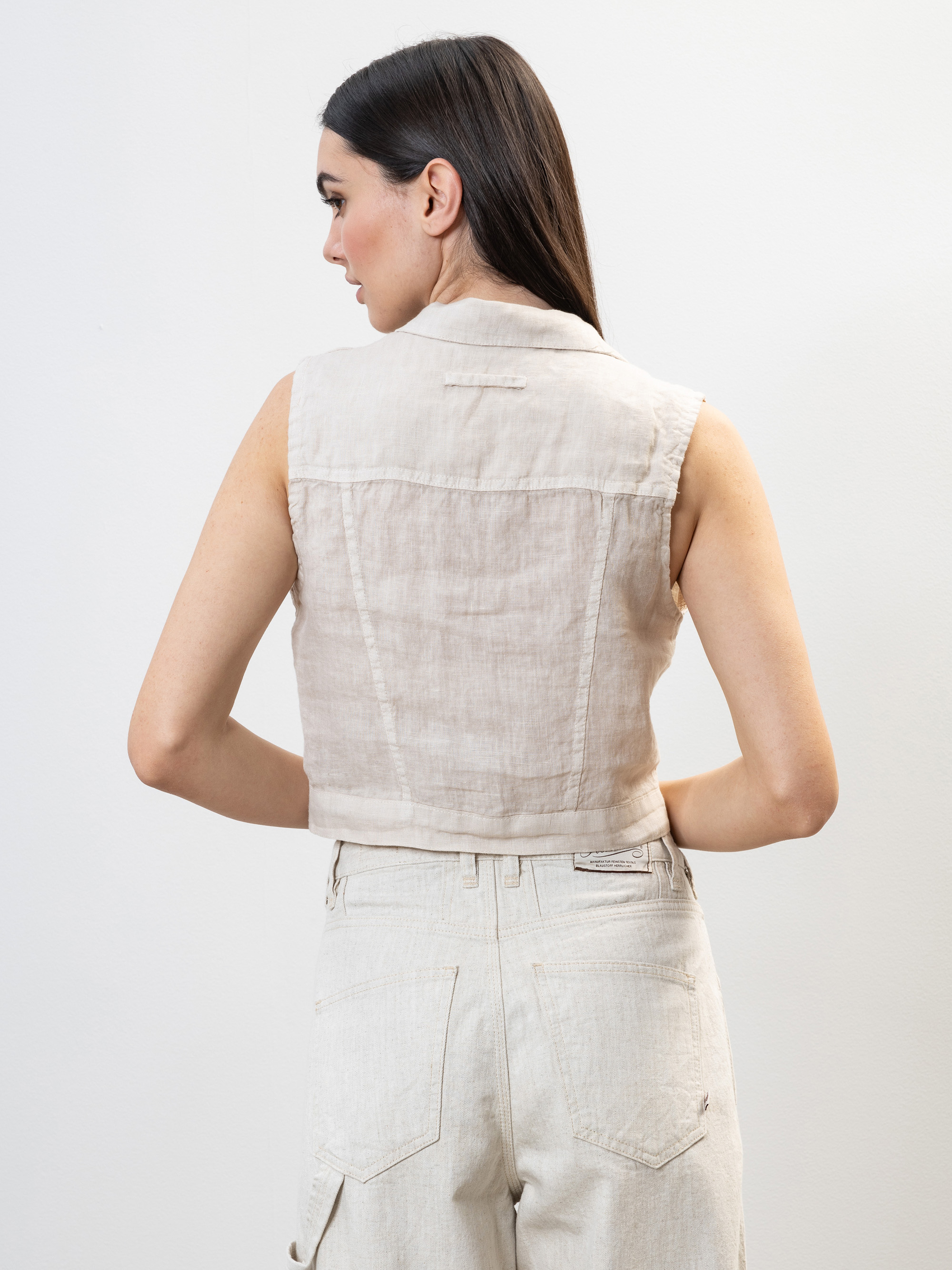 Eine Frau mit langen dunklen Haaren ist von hinten zu sehen. Sie trägt eine ärmellose beigefarbene Leinenweste und eine cremefarbene Jeans mit hoher Taille und steht vor einem schlichten hellen Hintergrund.