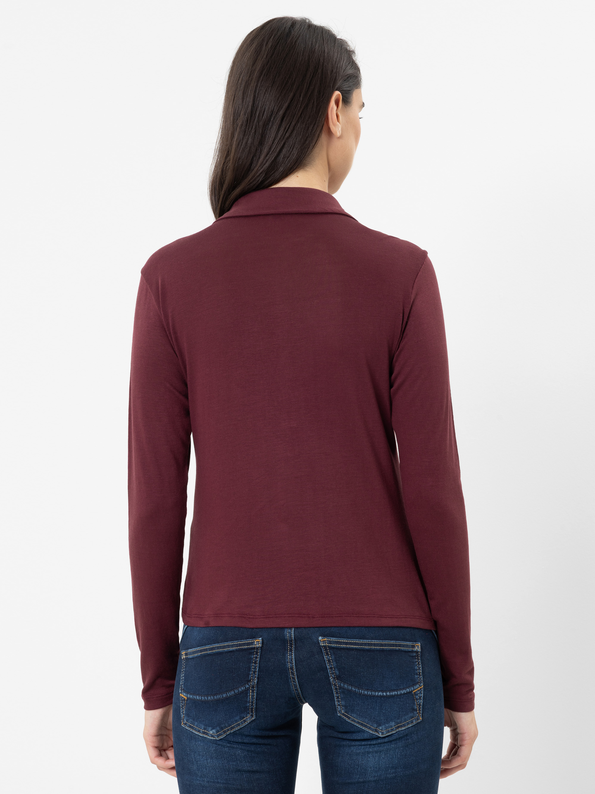 Eine Frau mit langen dunklen Haaren, die ein langärmeliges, weinrotes Oberteil und blaue Jeans trägt, ist vor einem schlichten weißen Hintergrund zu sehen, wobei sie mit dem Rücken zur Kamera steht.