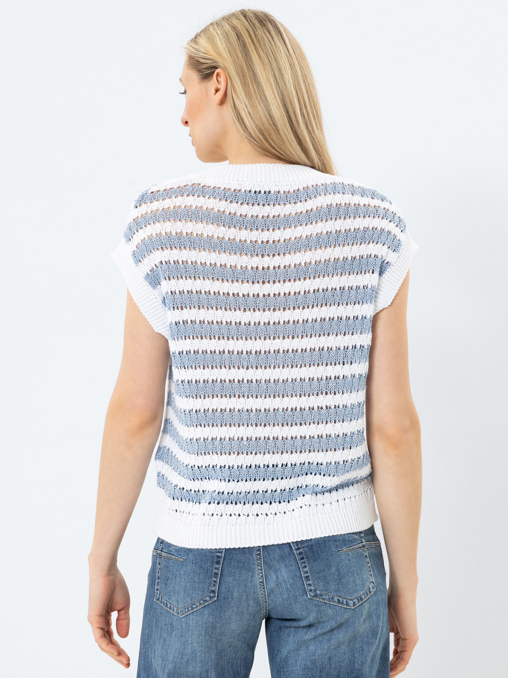 Eine Frau mit langen blonden Haaren ist von hinten zu sehen. Sie trägt ein kurzärmeliges, blau-weiß gestreiftes Strickoberteil mit weißem Saum und blaue Jeans vor einem schlichten hellen Hintergrund.