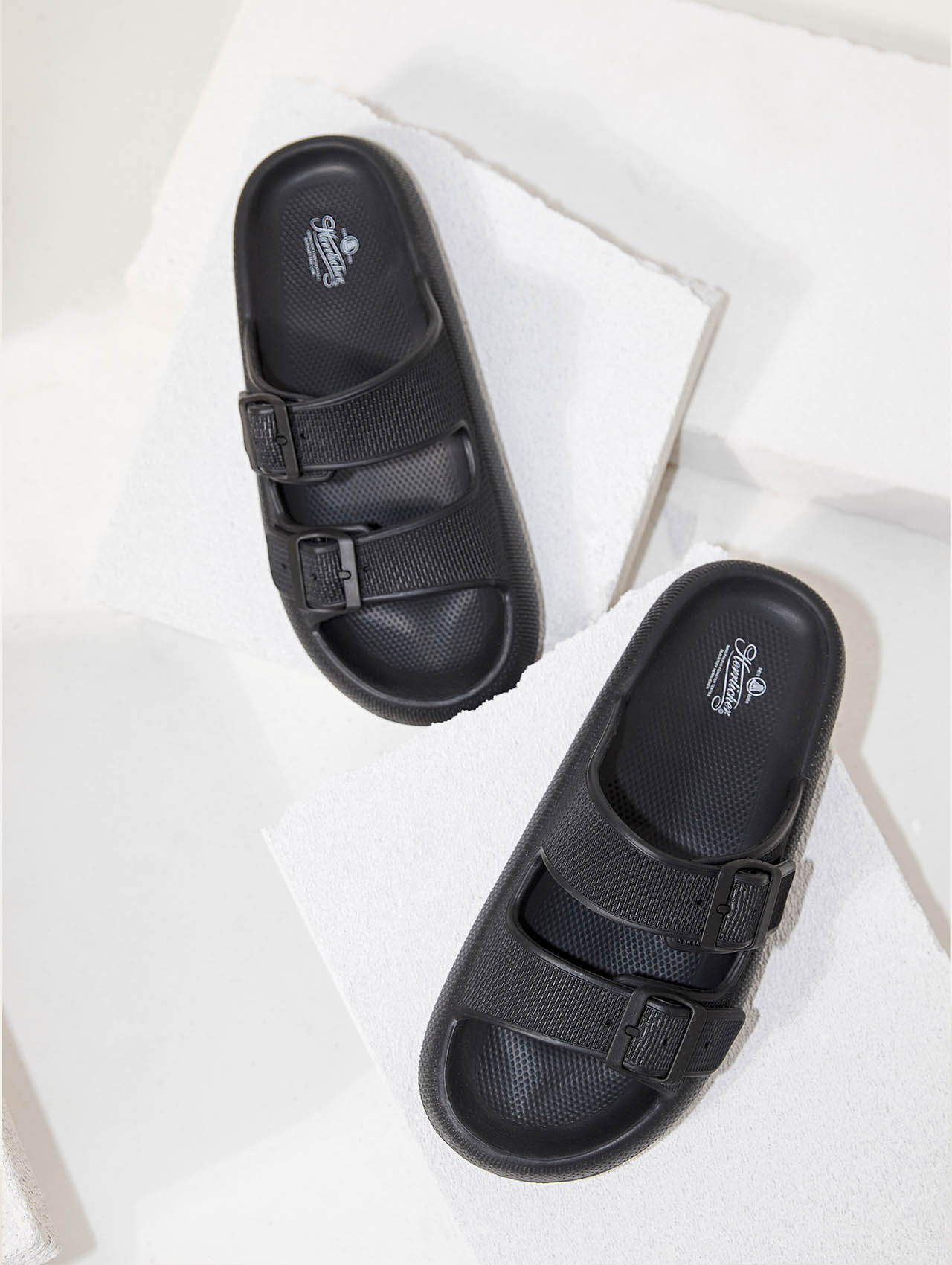 Ein Paar schwarzer, doppelreihiger Sandalen mit Schnallen ist auf weißen, strukturierten Blöcken abgebildet, von oben nach unten betrachtet.