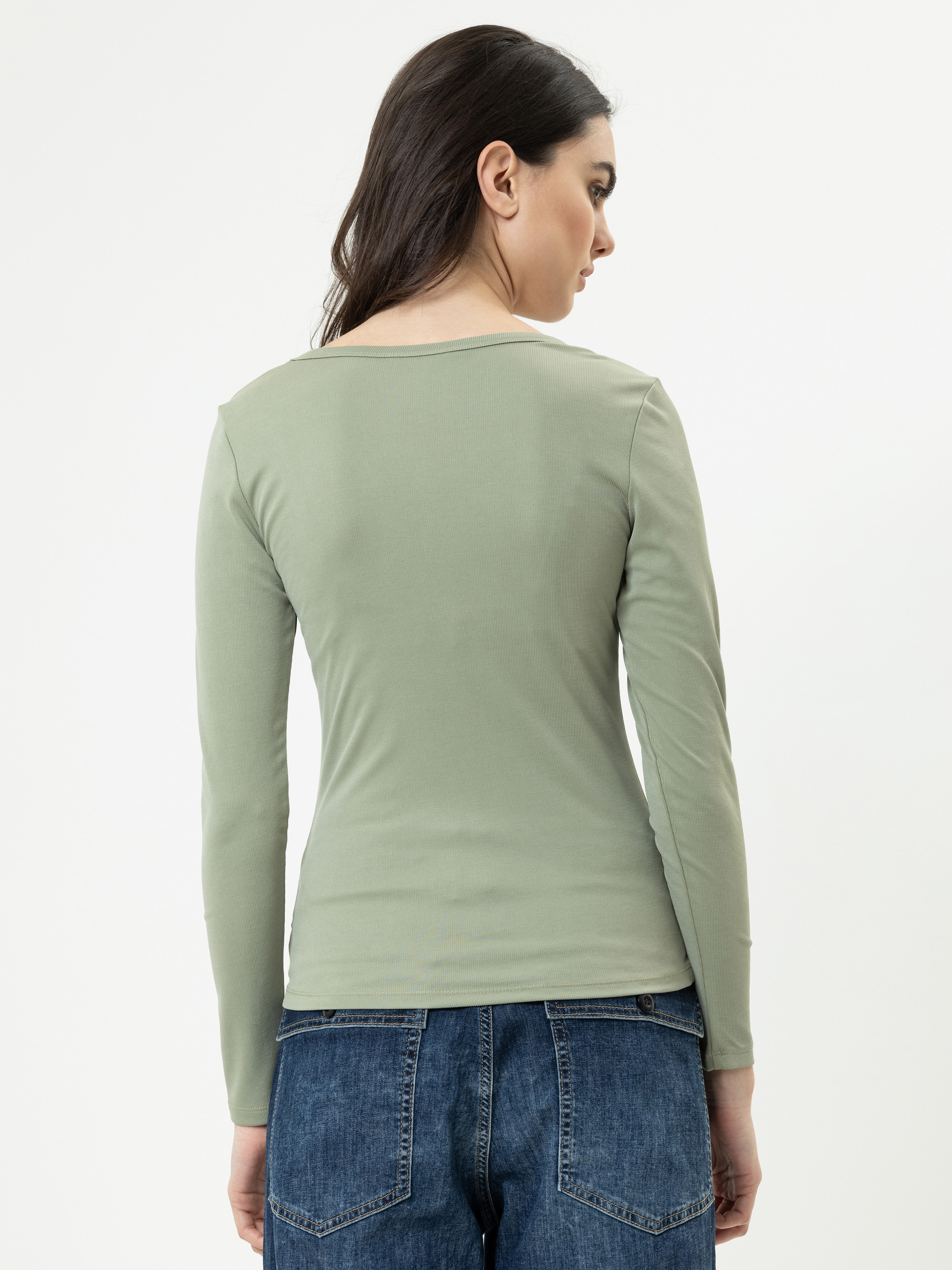 Eine Frau mit langen dunklen Haaren trägt ein hellgrünes langärmeliges Oberteil und blaue Jeans und steht mit dem Rücken zur Kamera vor einem schlichten weißen Hintergrund.