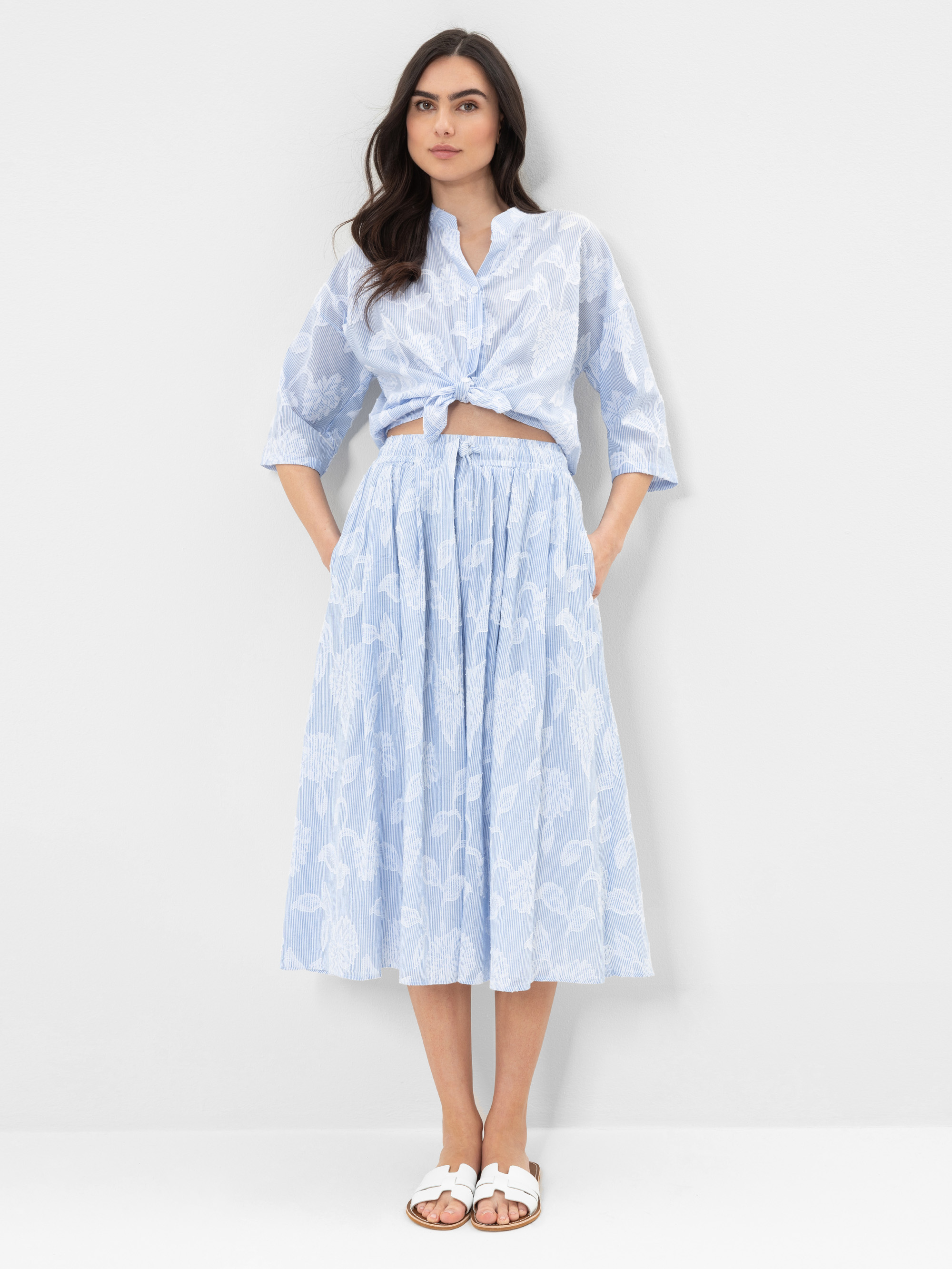 Eine Frau steht vor einem schlichten weißen Hintergrund und trägt eine hellblaue, durchsichtige Bluse mit Blumenmuster, die in der Taille gebunden wird, und einen passenden Midirock. Sie trägt weiße Sandalen und hat die Hände in den Taschen ihres Roc