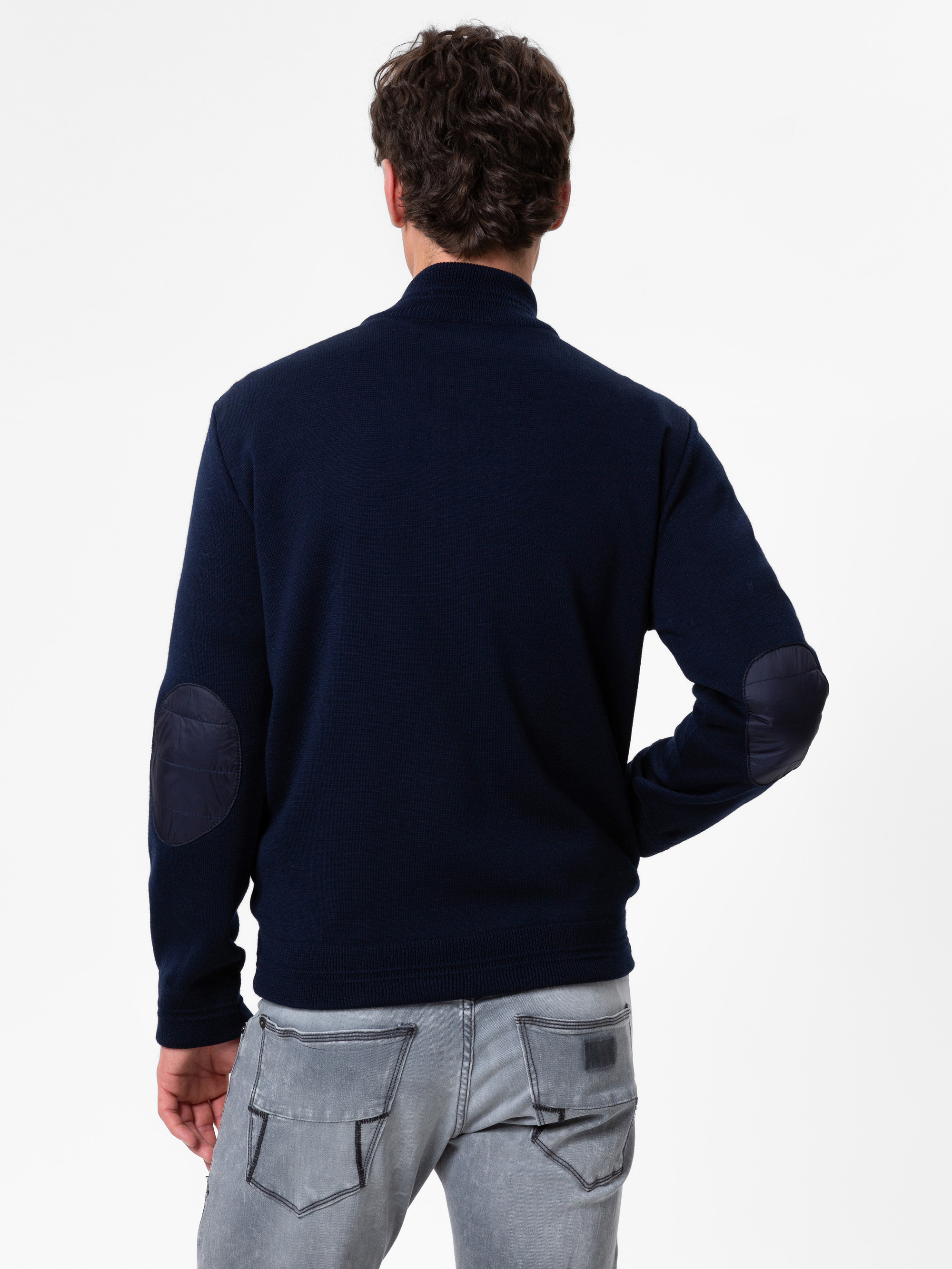 Ein Mann, der einen marineblauen Pullover mit dunklen Ellbogenaufnähern und hellgraue Jeans trägt, steht mit dem Rücken zur Kamera vor einem schlichten weißen Hintergrund.