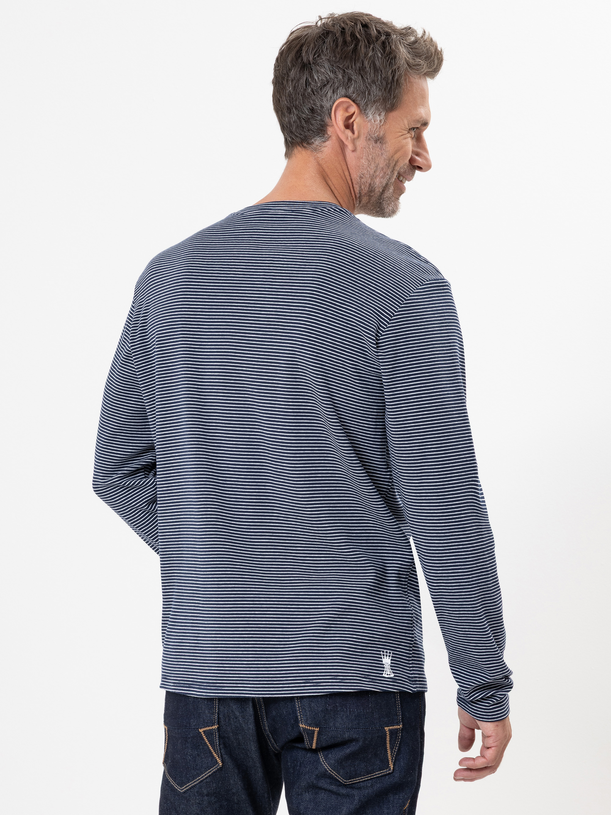 Ein Mann mit kurzen braunen Haaren und Bart blickt von sich weg und trägt ein langärmeliges, marineblau-weiß gestreiftes Hemd und dunkle Jeans vor einem schlichten weißen Hintergrund.