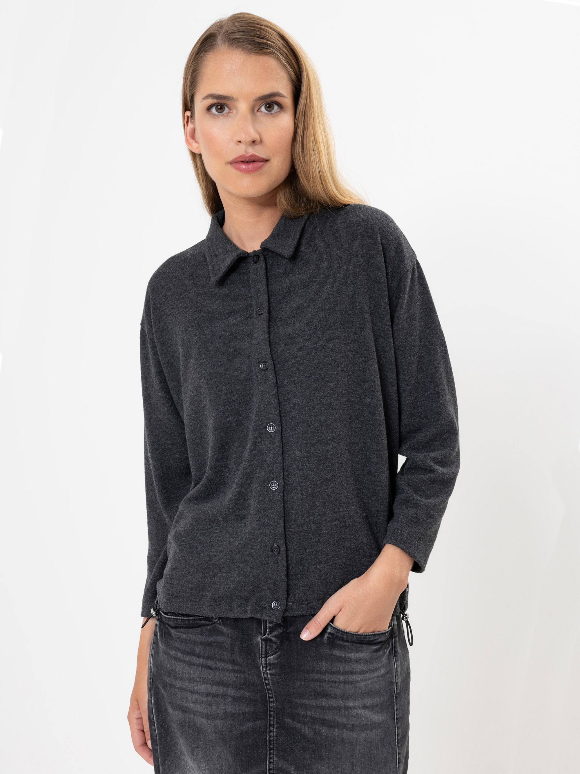 Eine Frau mit langen hellbraunen Haaren, die ein dunkelgraues Button-up-Hemd und eine schwarze, verwaschene Jeans trägt, steht vor einem schlichten weißen Hintergrund und schaut mit neutralem Blick direkt in die Kamera.