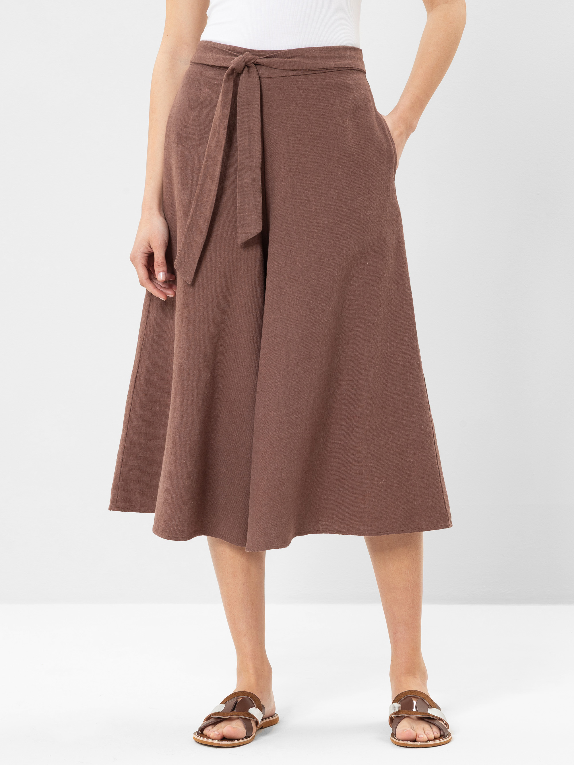 Eine Person, die eine hochtaillierte braune Hose mit weitem Bein und einem Stoffgürtel, ein weißes ärmelloses Oberteil und braune flache Sandalen trägt, steht vor einem schlichten hellen Hintergrund.