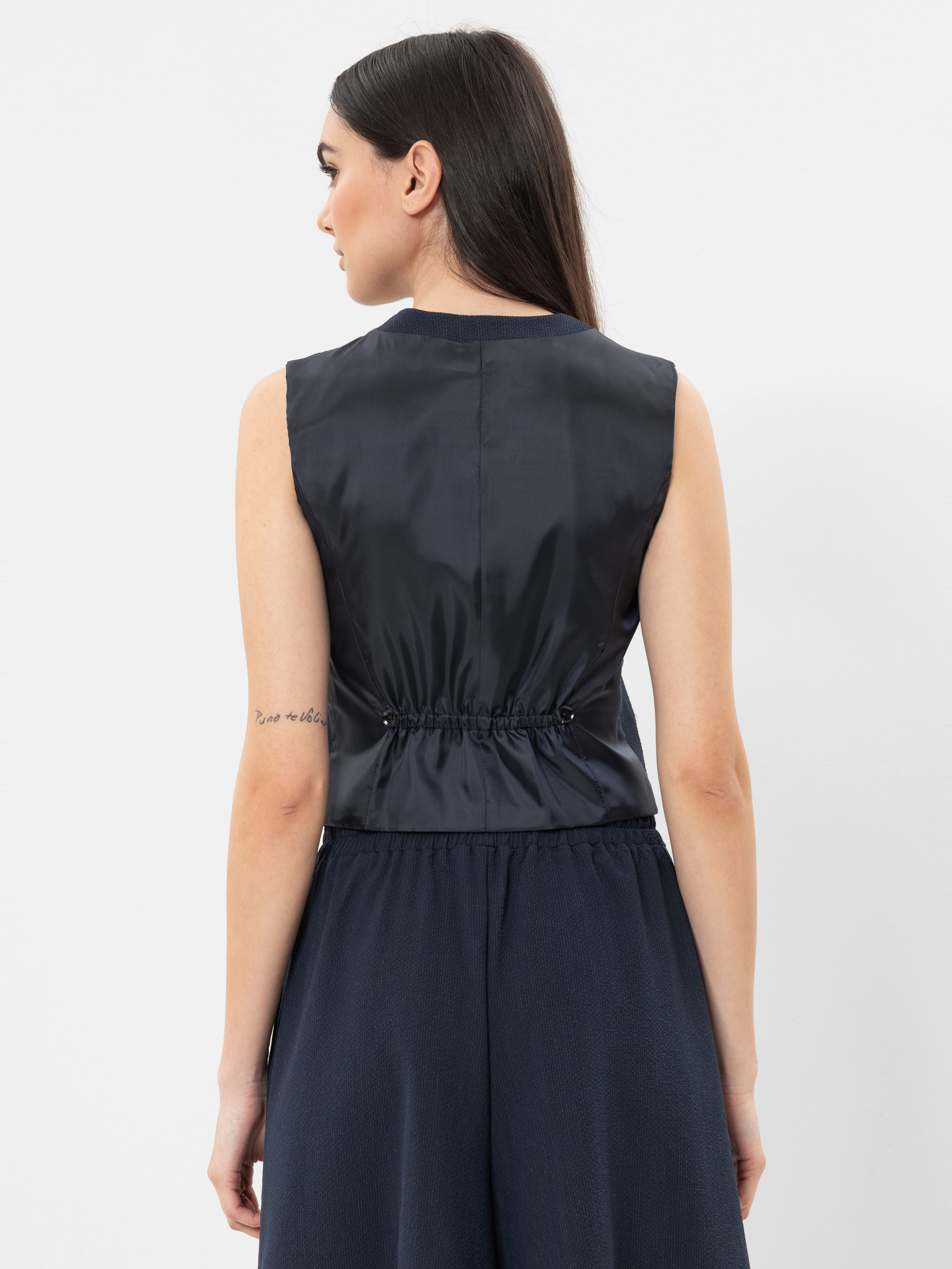 Eine Frau mit langem, glattem dunklem Haar ist von hinten zu sehen. Sie trägt ein ärmelloses, marineblaues Oberteil mit enger Taille und eine dazu passende Hose mit weitem Bein. Sie hat eine kleine Tätowierung auf ihrem rechten Unterarm. Der Hintergrun
