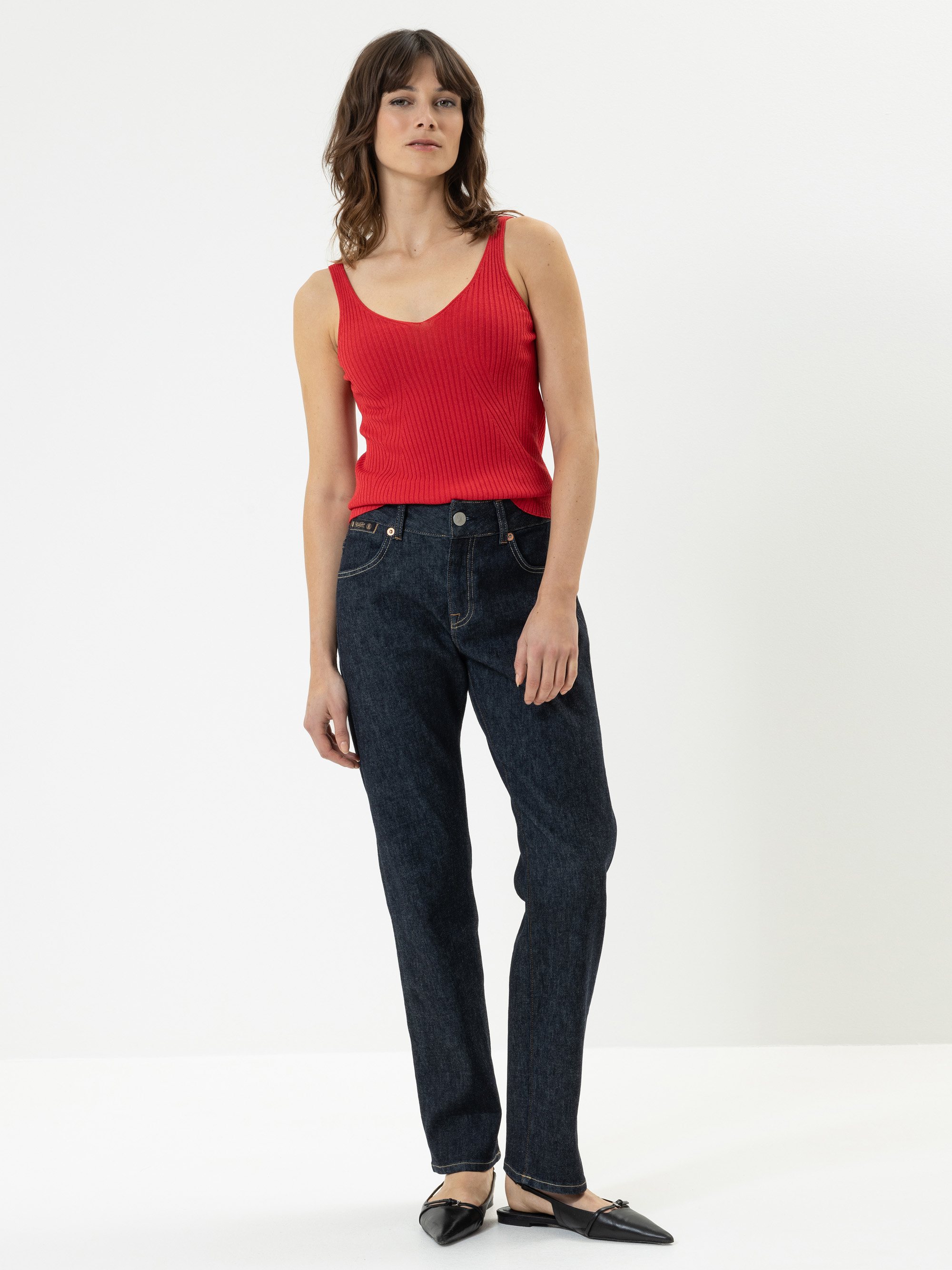Eine Frau steht vor einem weißen Hintergrund und trägt ein rotes ärmelloses Oberteil, dunkelblaue Jeans und schwarze flache Schuhe. Sie hat schulterlanges braunes Haar und einen neutralen Gesichtsausdruck.