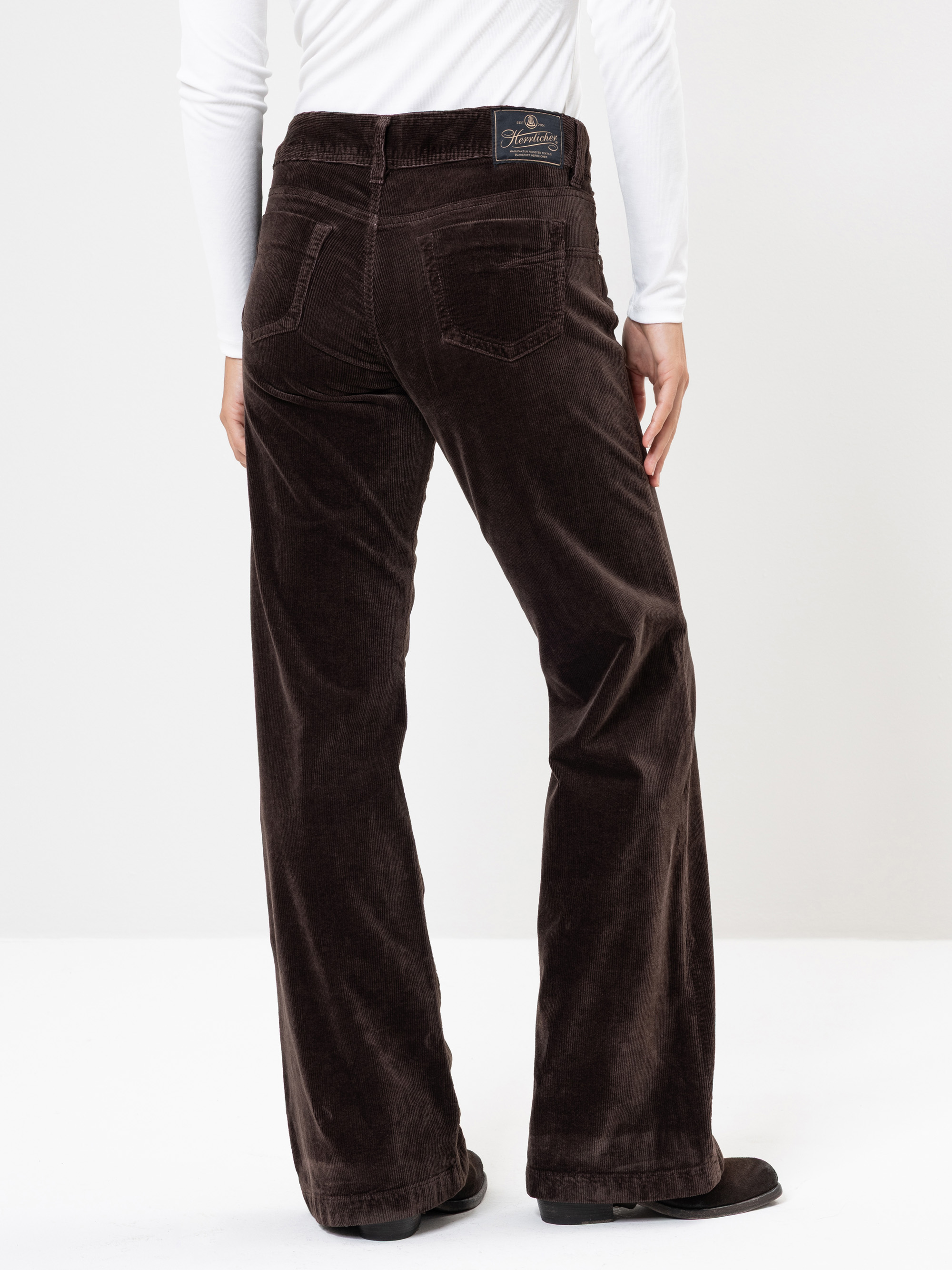 Eine Person, die eine dunkelbraune Cordhose und ein weißes langärmeliges Hemd trägt, wird von hinten gezeigt und steht vor einem schlichten weißen Hintergrund.