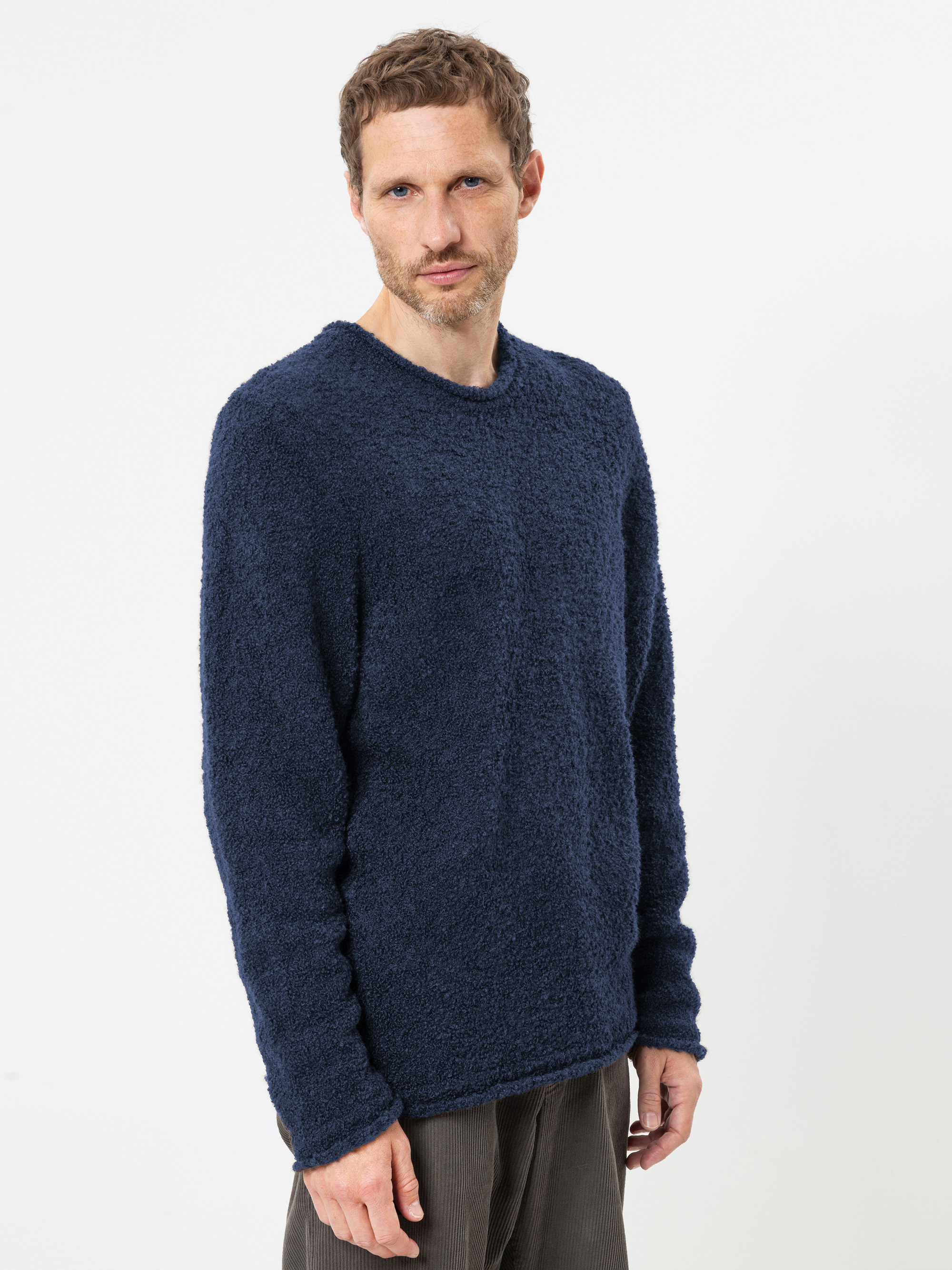 Ein Mann mit kurzen braunen Haaren und einem gepflegten Bart trägt einen strukturierten marineblauen Pullover und eine dunkle Hose. Er steht vor einem schlichten weißen Hintergrund und blickt mit neutralem Gesichtsausdruck leicht zur Seite.
