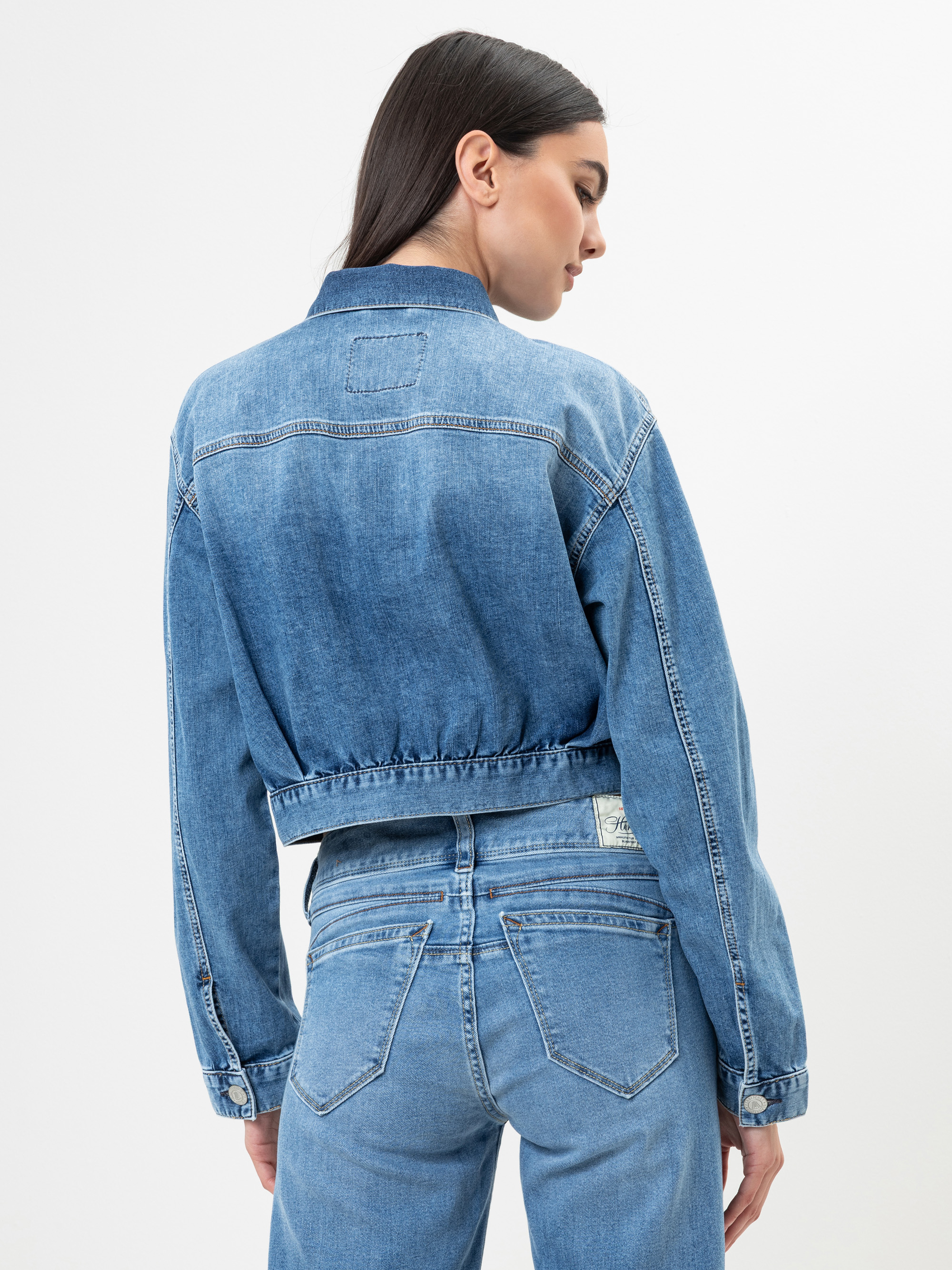 Eine Frau mit langen dunklen Haaren, die eine abgeschnittene blaue Jeansjacke und hochgekrempelte Bluejeans trägt, steht mit dem Gesicht zur Seite und blickt über ihre Schulter auf einen schlichten weißen Hintergrund.