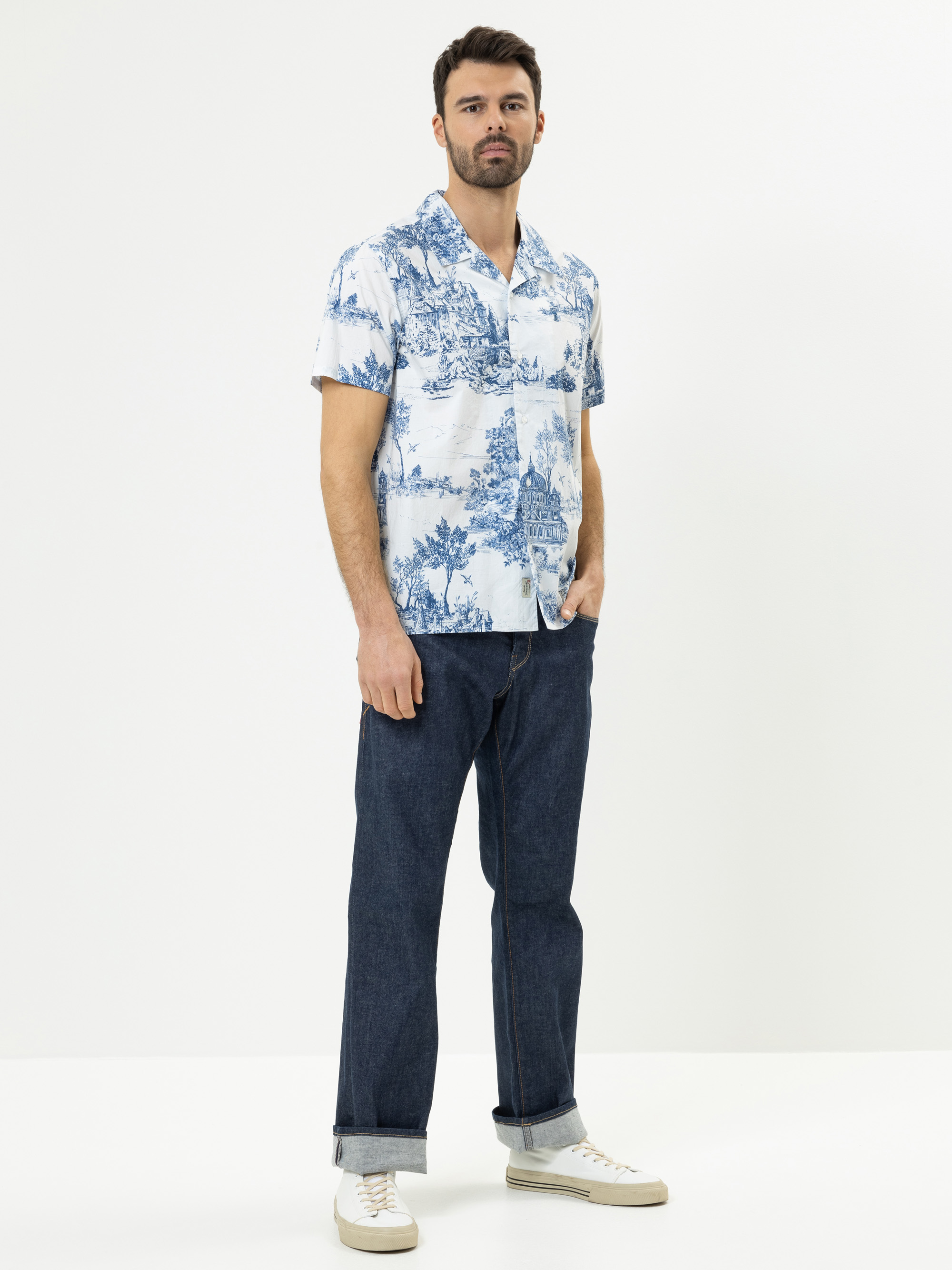 Ein Mann steht vor einem schlichten weißen Hintergrund und trägt ein blau-weiß gemustertes kurzärmeliges Hemd, dunkelblaue Jeans mit Bündchen und weiße Turnschuhe. Er hat kurzes dunkles Haar, einen Bart und einen entspannten Gesichtsausdruck.