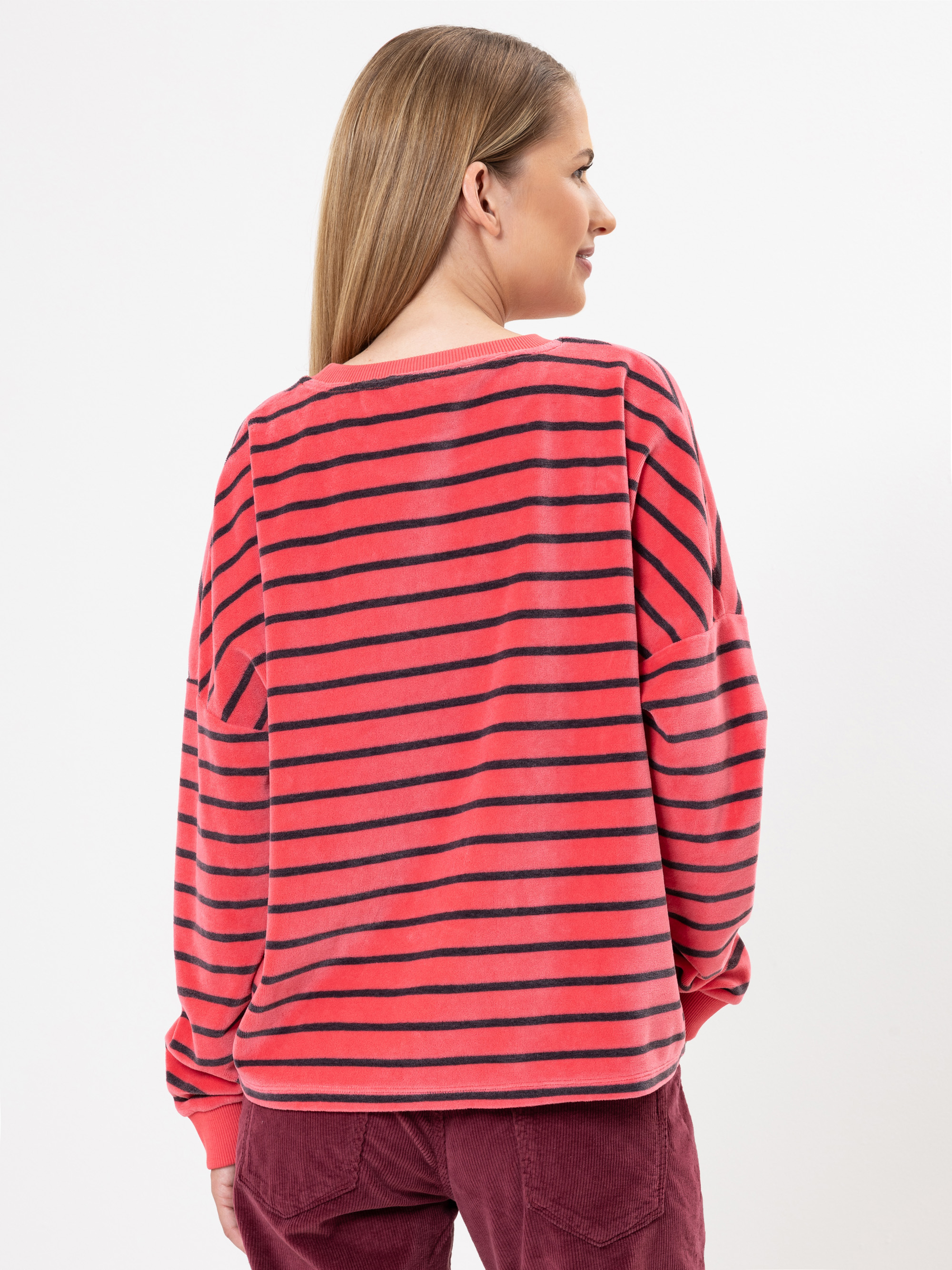 Eine Frau mit glattem, hellbraunem Haar trägt ein rot-schwarz gestreiftes, langärmeliges Hemd und eine kastanienbraune Hose und steht mit dem Rücken zur Kamera vor einem schlichten weißen Hintergrund.